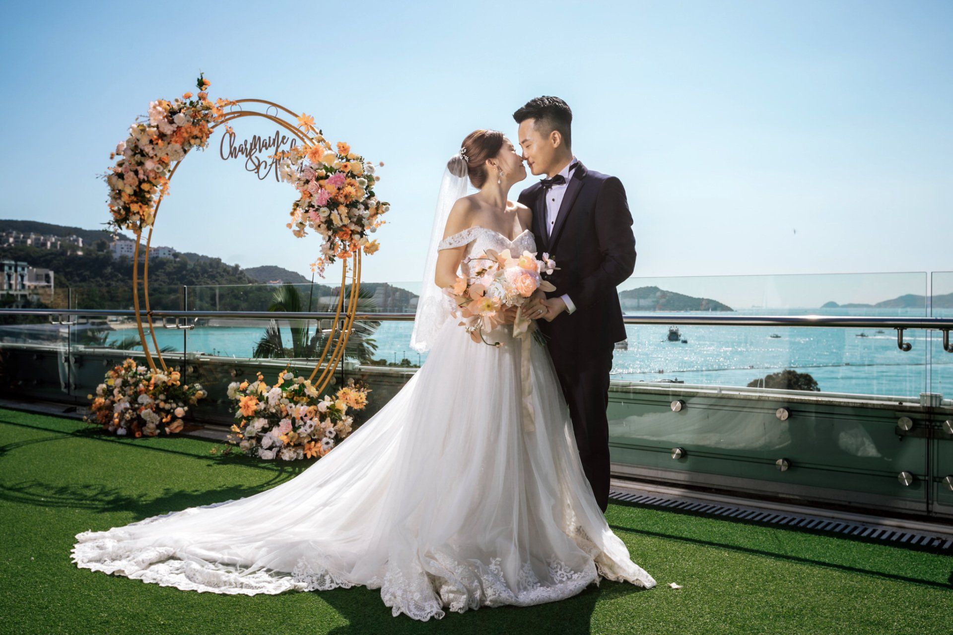 Bride and groom embrace by floral arch overlooking ocean.