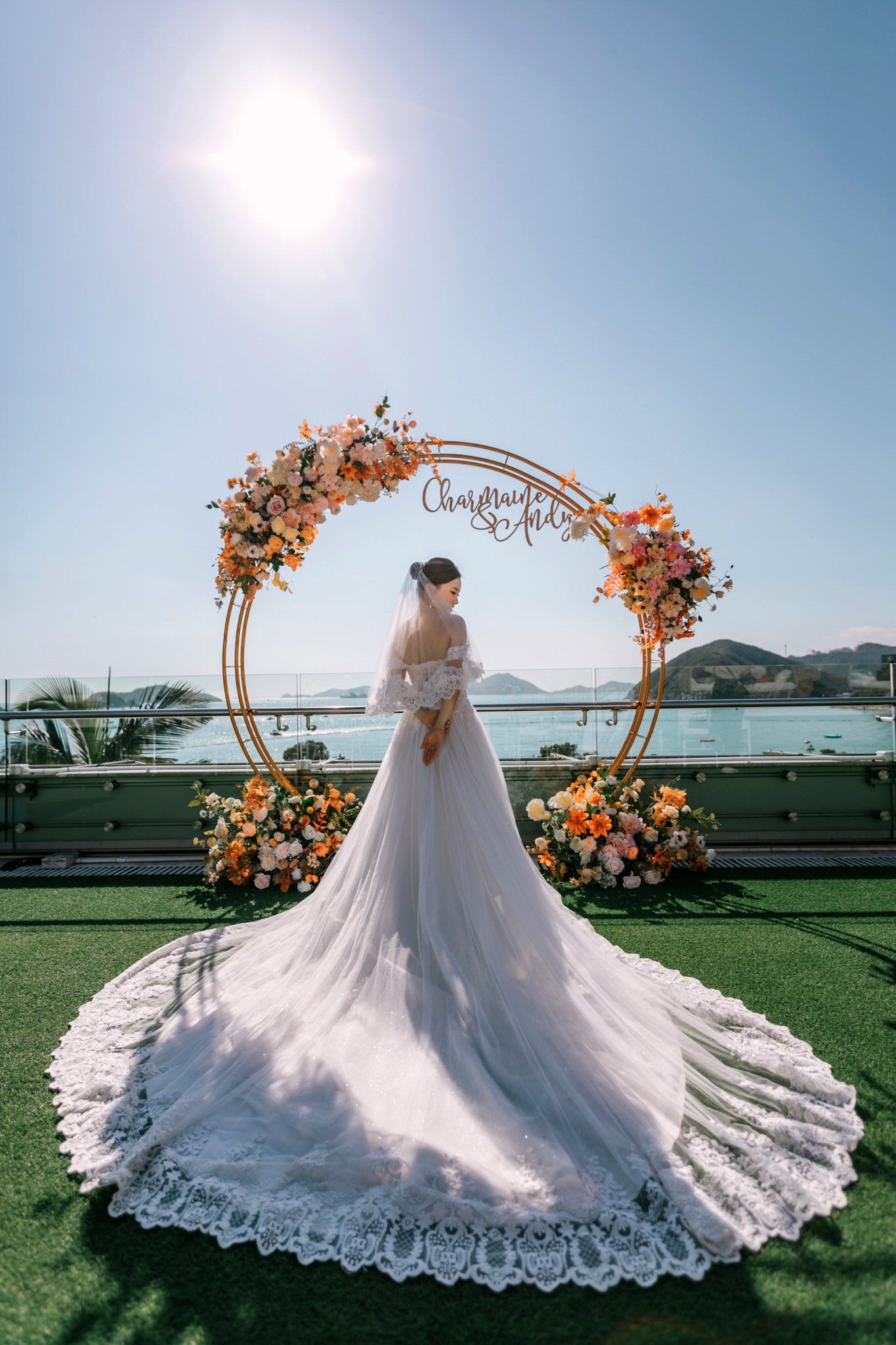Bride in white gown stands before floral arch on rooftop overlooking water.