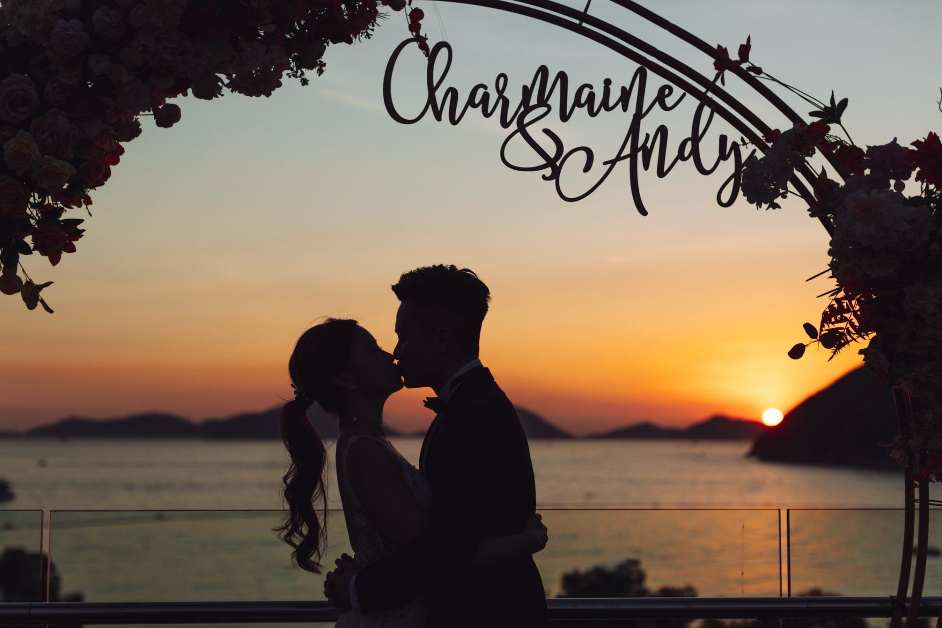 Couple kissing at sunset under an arch with floral accents and names 