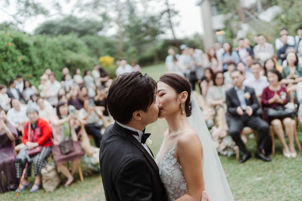 Bride and groom kissing at outdoor wedding ceremony; crowd seated in background.