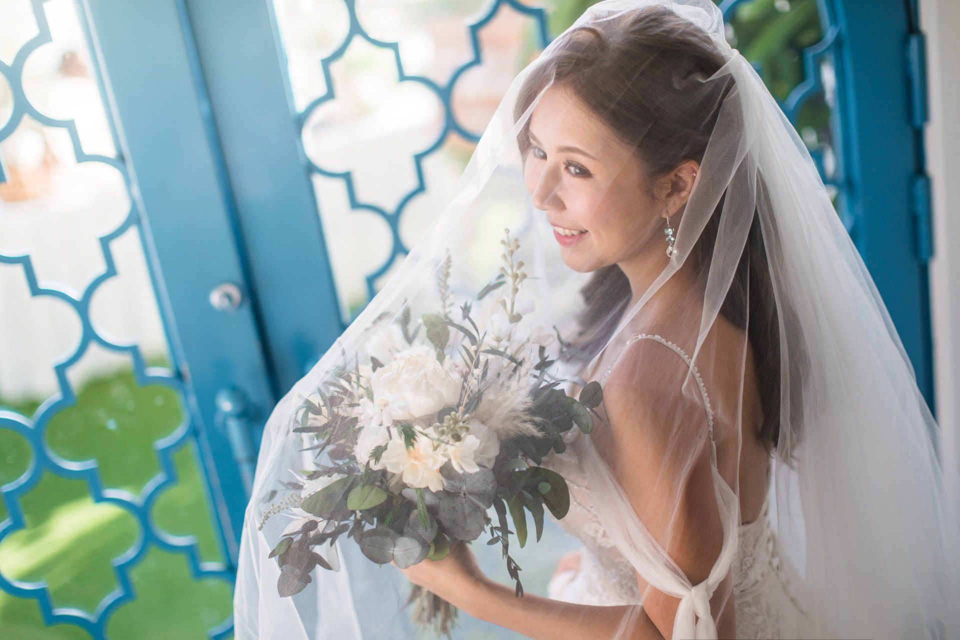 Bride in a veil holding a bouquet, standing near a blue door, looking off to the side, smiling.