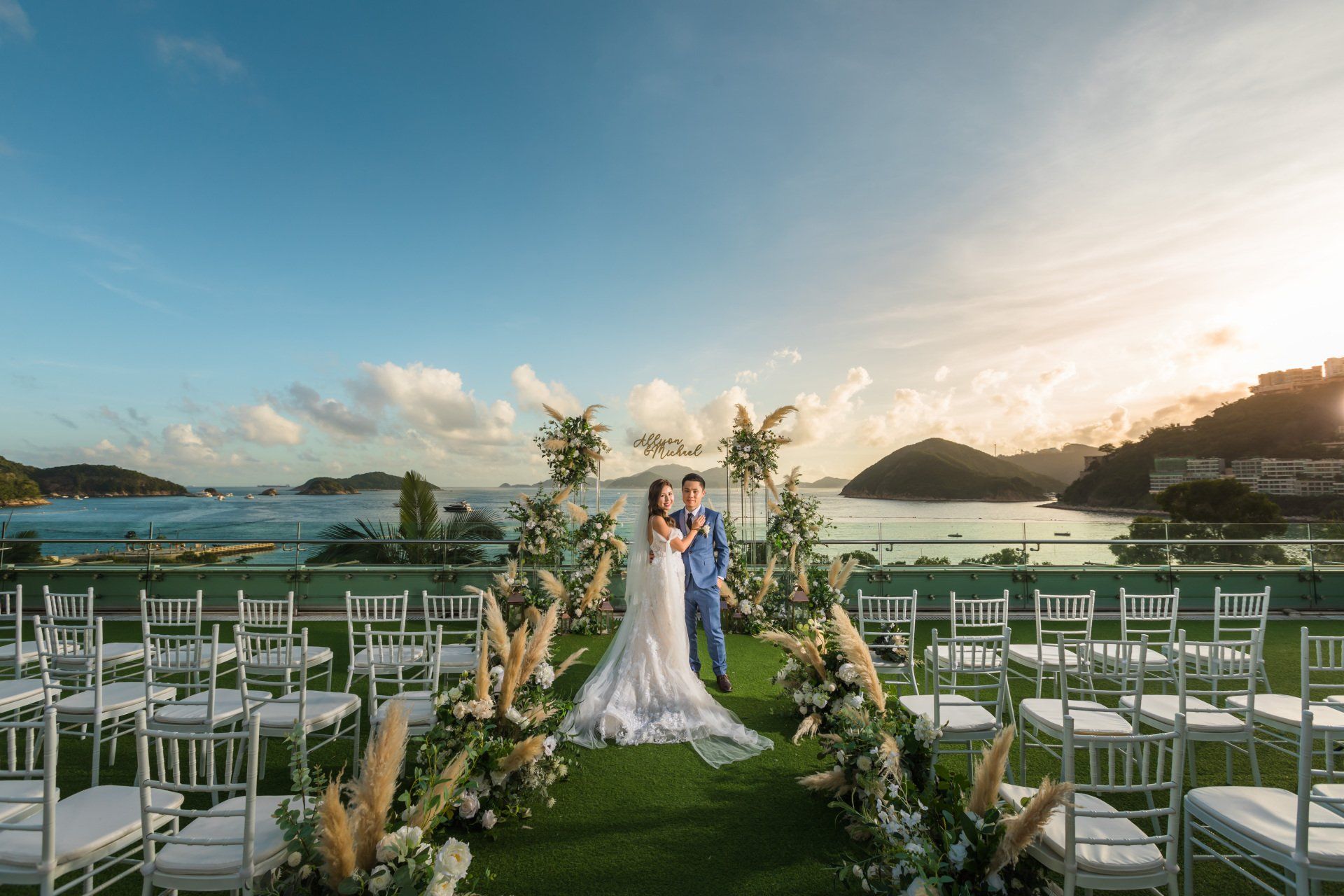 Couple at outdoor wedding ceremony; ocean backdrop, archway with flowers, white chairs.