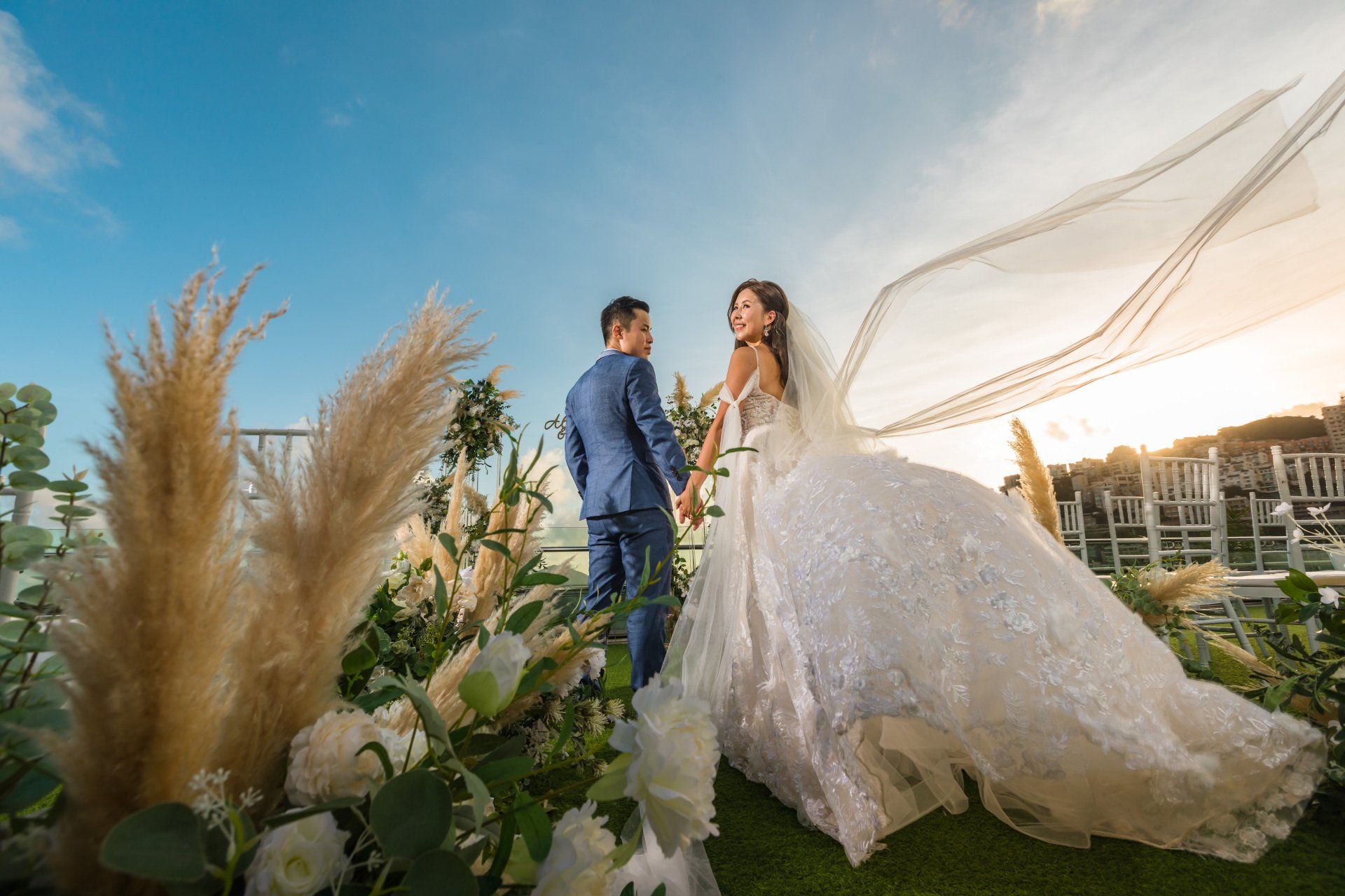 Couple in wedding attire walk outdoors, bride's veil blowing in the wind, holding hands, sunny day.
