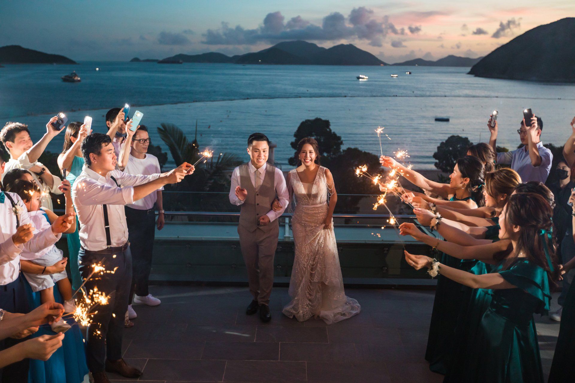 Newlyweds run through a sparkler-lined pathway, with ocean backdrop.