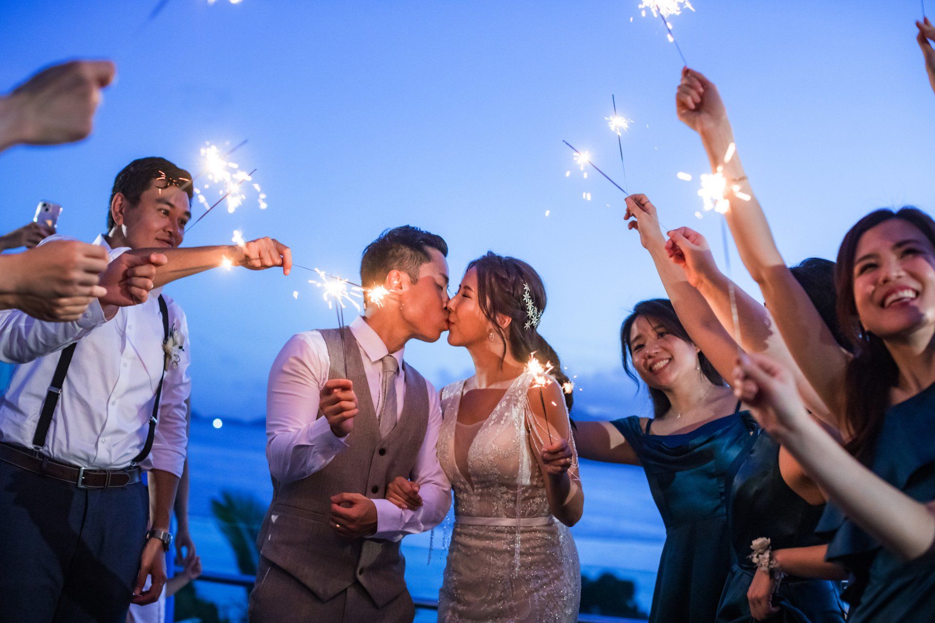 Newlyweds kiss amidst sparklers held by wedding guests at dusk.