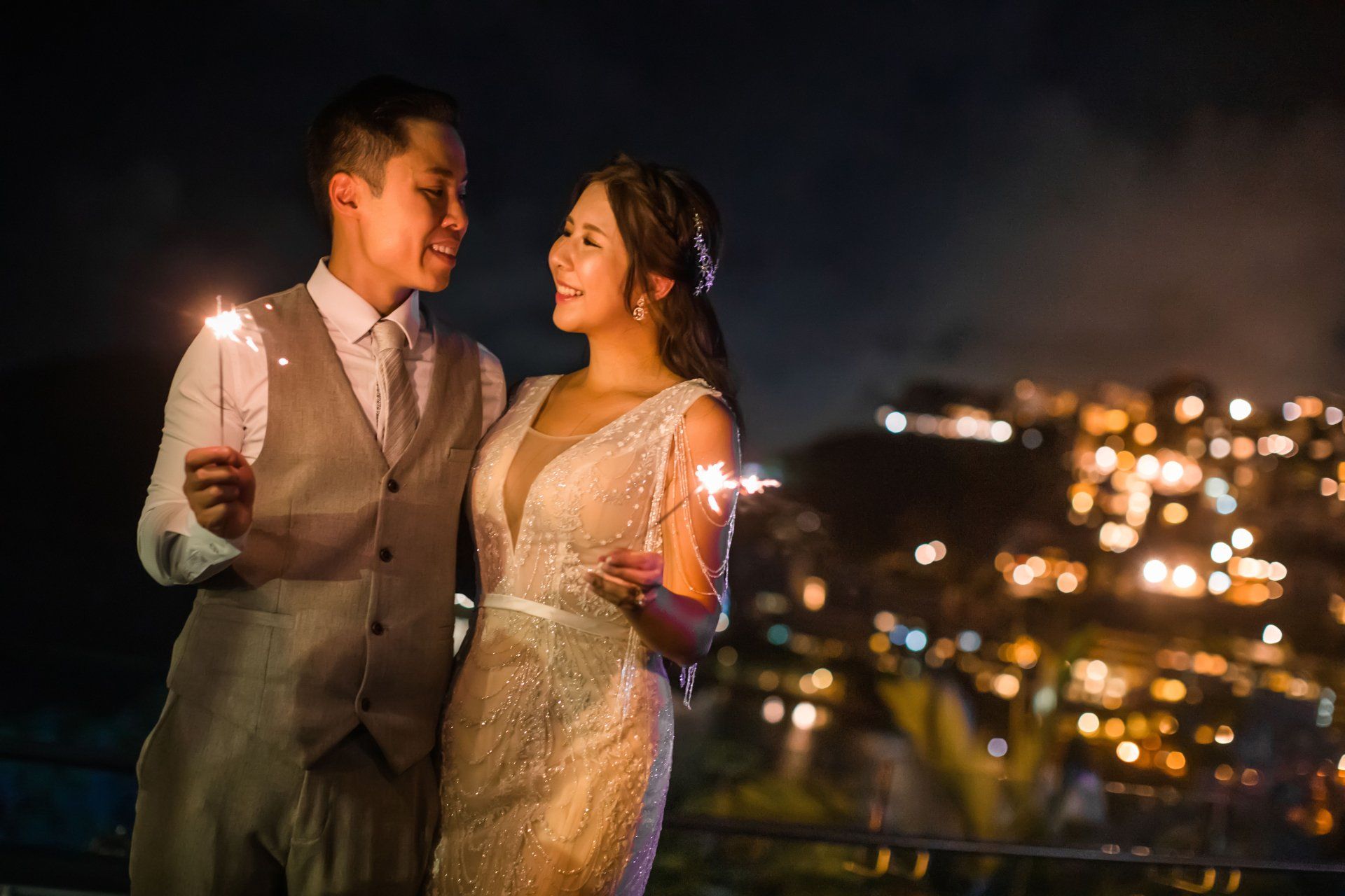 Couple holding sparklers, gazing at each other on a balcony at night, overlooking city lights.