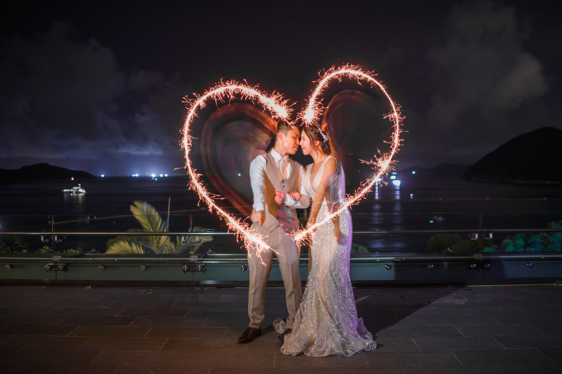 Couple kissing in front of a sparkler heart at night, overlooking a harbor.