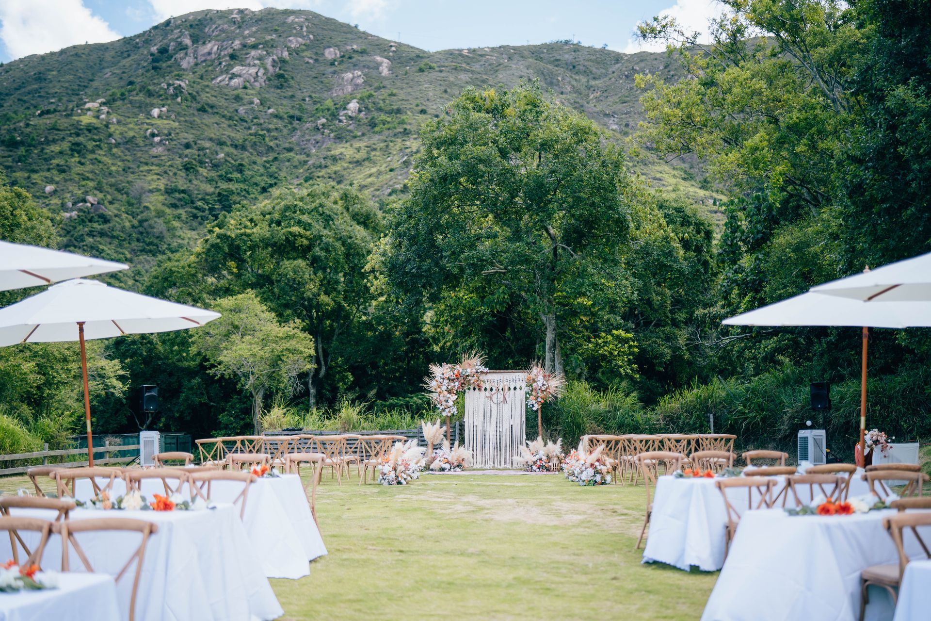 Wedding ceremony setup in a grassy outdoor area with mountain backdrop, white umbrellas, and wooden chairs.
