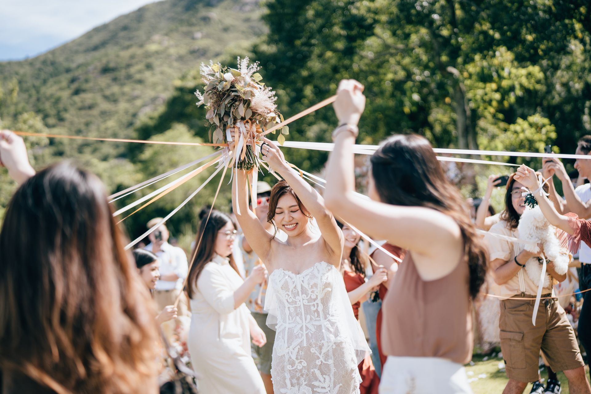 Bride tossing bouquet amidst ribbon dance, smiling outdoors. People in background.