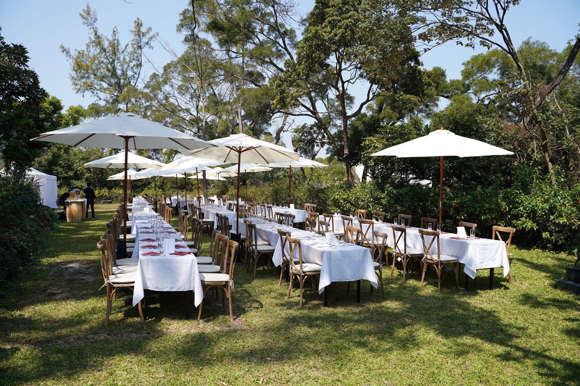 Outdoor event with long tables covered in white linens, under umbrellas, set on grass.