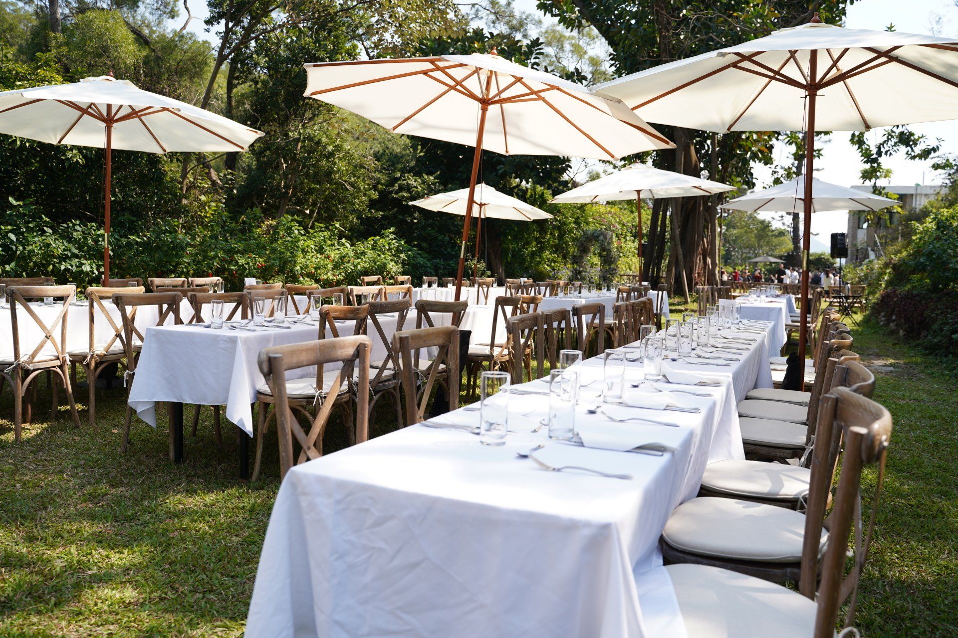 Outdoor dining tables set with white tablecloths, chairs, and umbrellas in a garden setting.