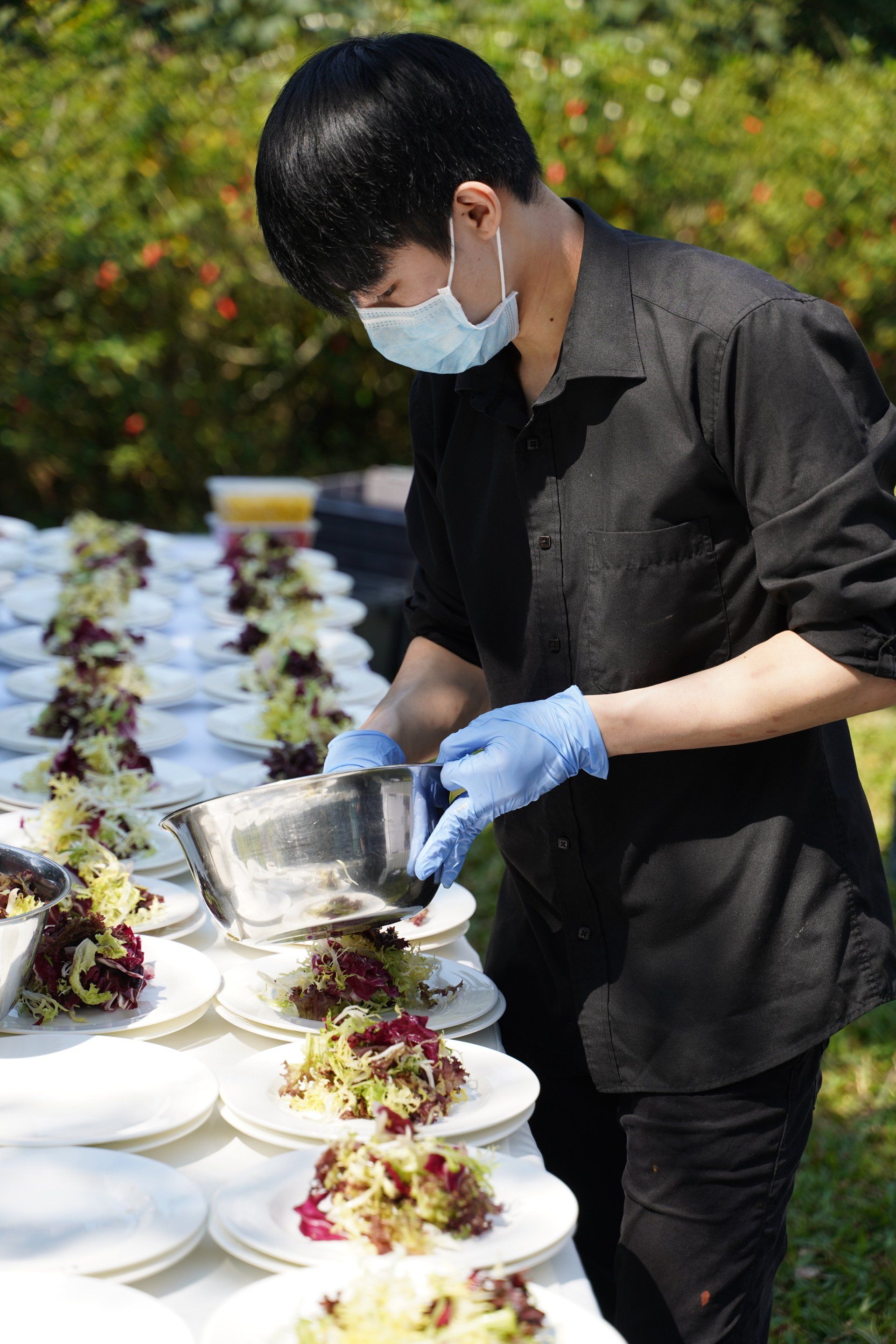 Person in black shirt and mask, adding dressing to salads on white plates, outdoors.