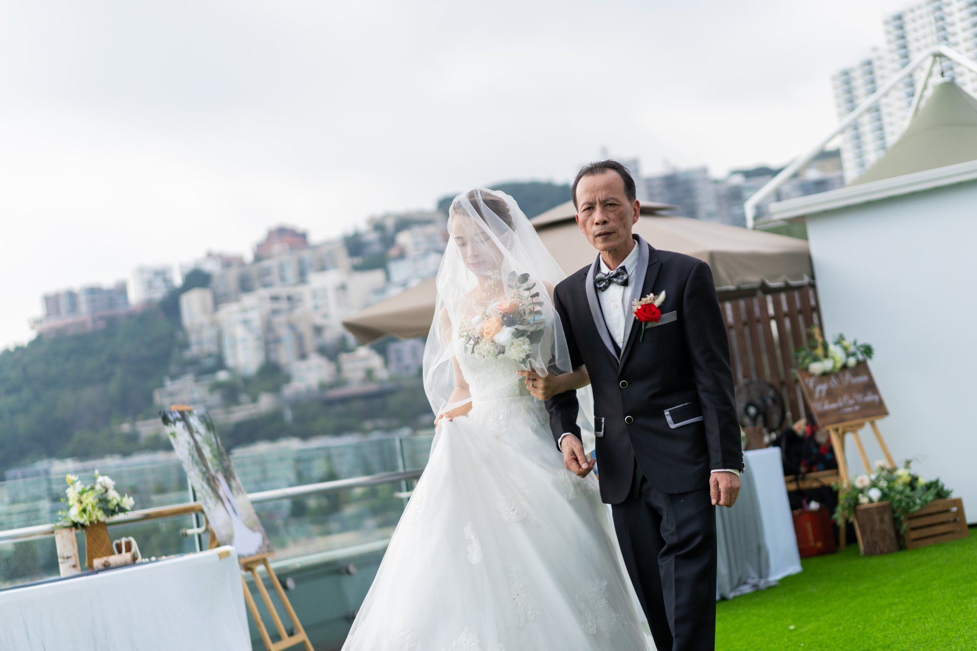Bride in a white gown and veil walks with her father down an outdoor aisle. City in background.