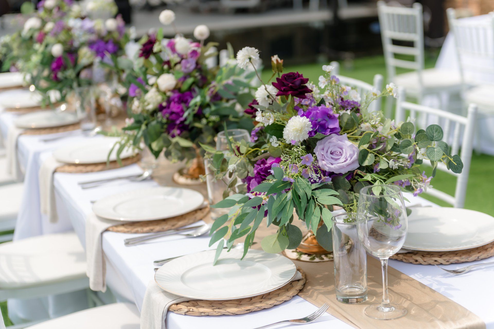 Formal table setting with purple floral centerpieces, white plates, and champagne glasses outdoors.