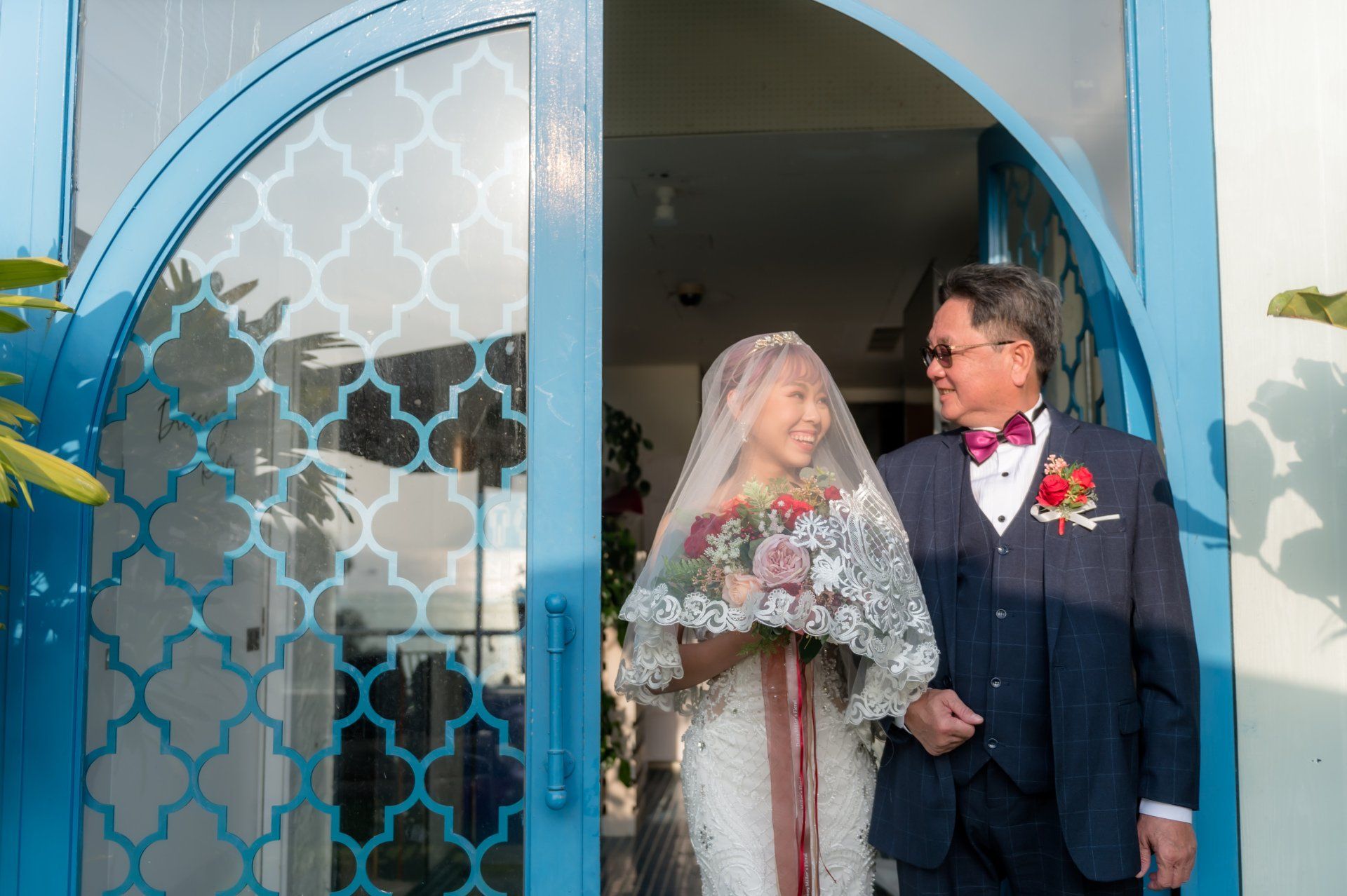 Bride and man in formal attire exiting a blue-arched doorway. Bride holds a bouquet and wears a veil.