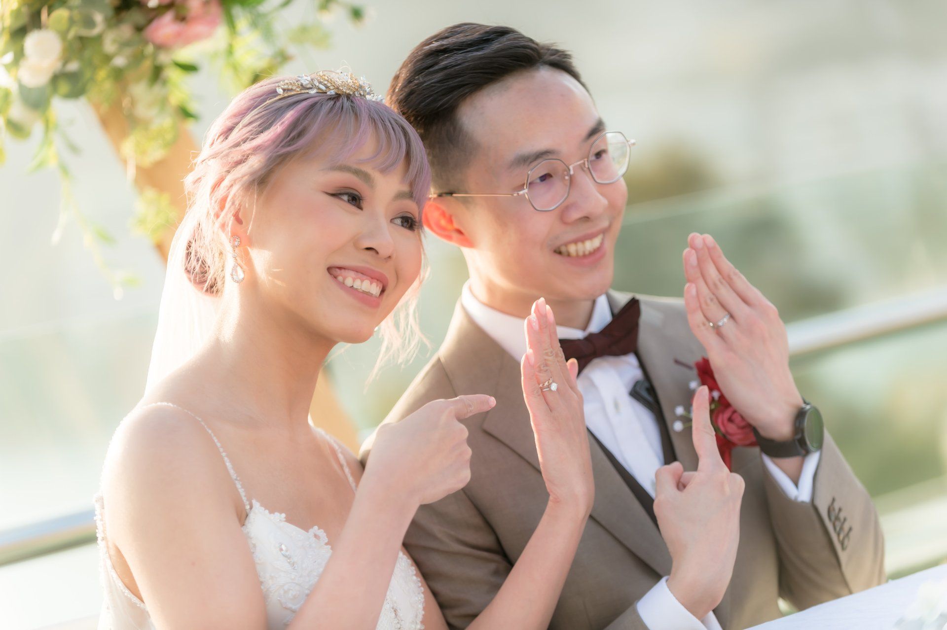 Newlyweds showing off wedding rings, smiling. Bride has lavender hair, wearing a tiara. Groom wears glasses and bow tie.