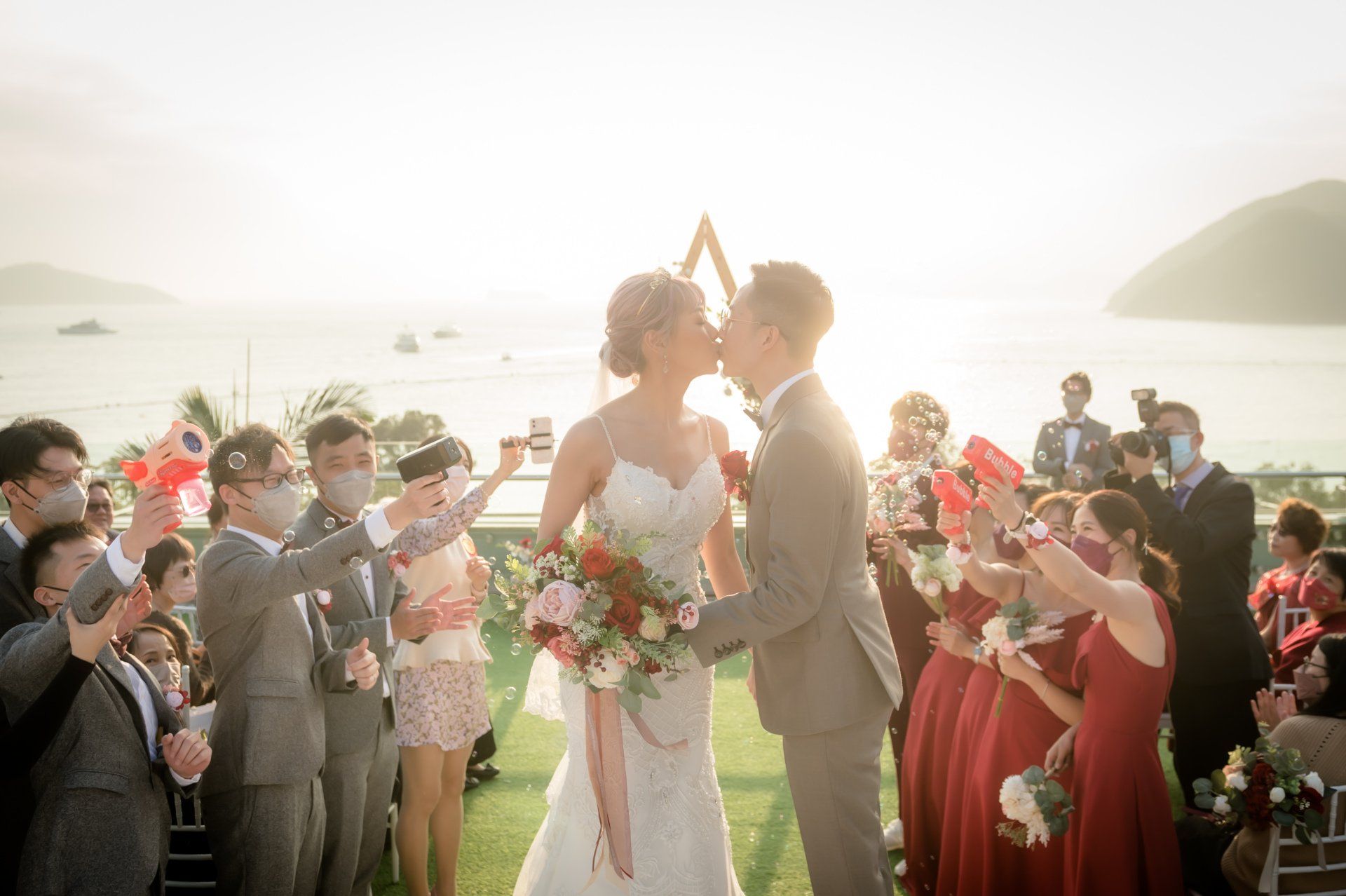 Couple kissing at outdoor wedding; guests cheering with confetti; ocean view.