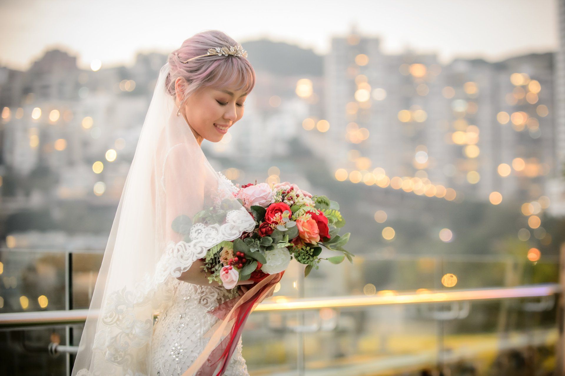 Bride in a wedding dress, holding flowers, smiling, with city lights blurred in the background.