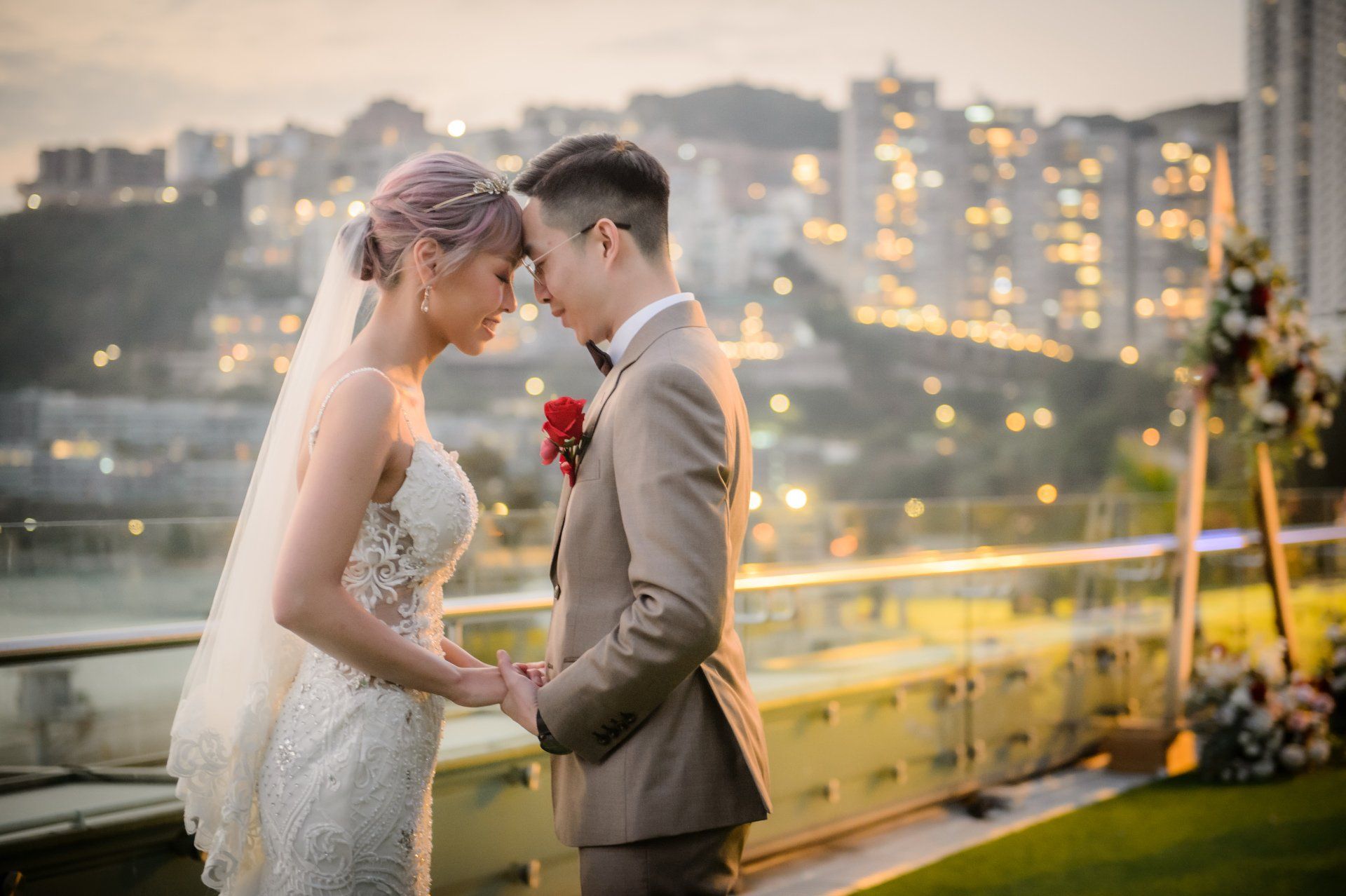 Bride and groom touch foreheads, holding hands. Rooftop ceremony with city lights in the background.