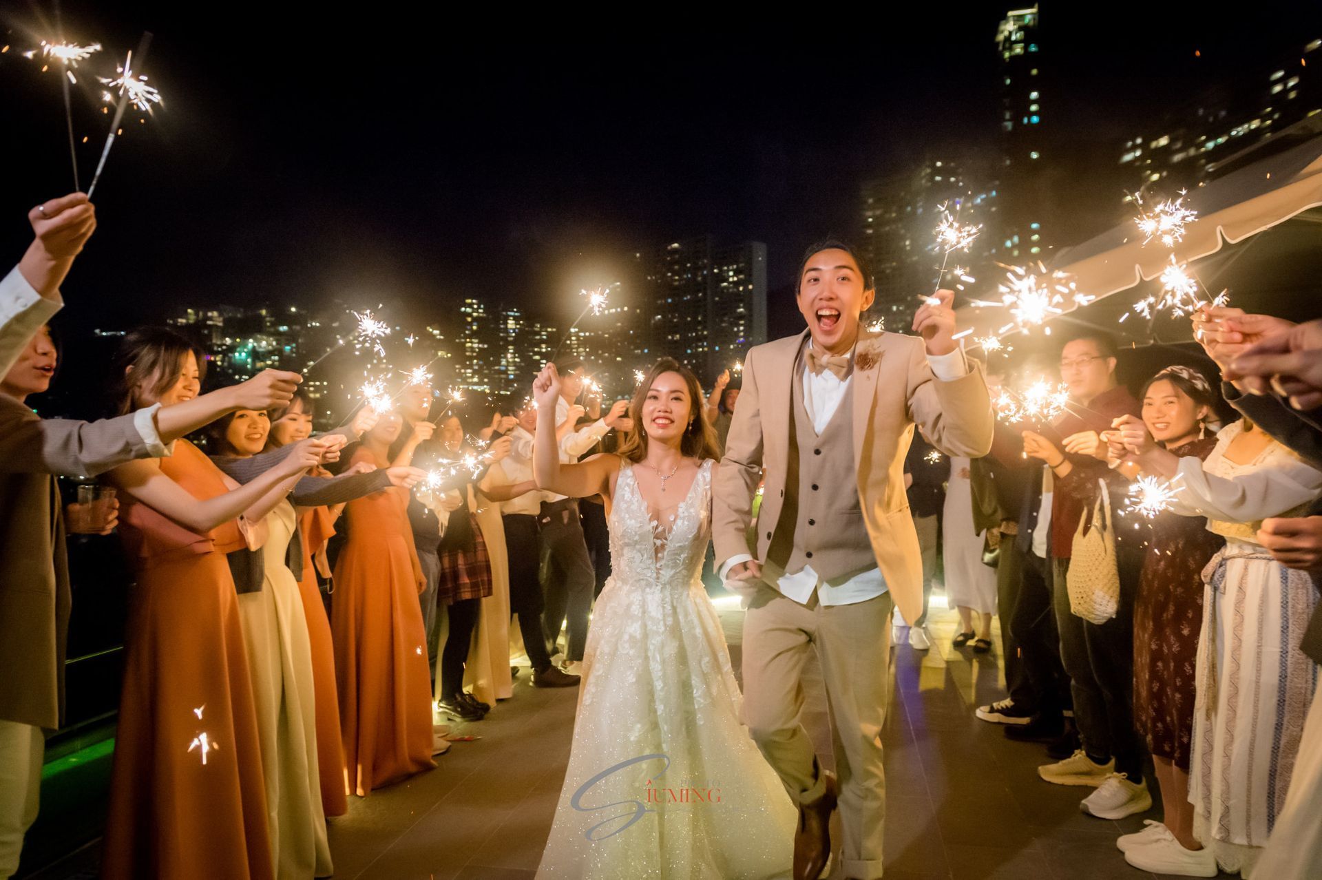 Newlyweds running through a sparkler-lit tunnel, smiling, at an outdoor evening wedding.