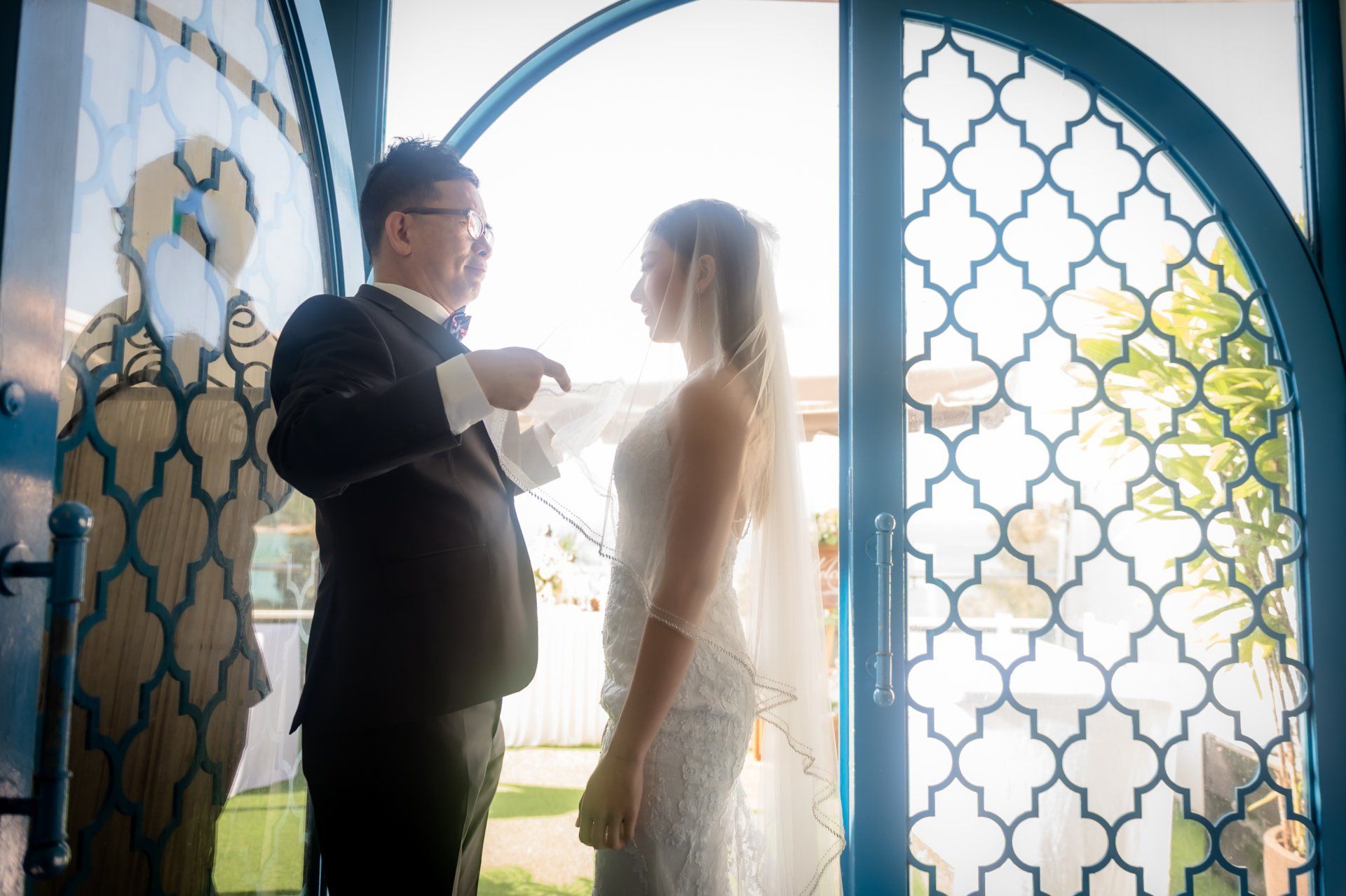 Groom in tuxedo adjusts bride's attire, standing in a doorway; sunlight streams through the open door.