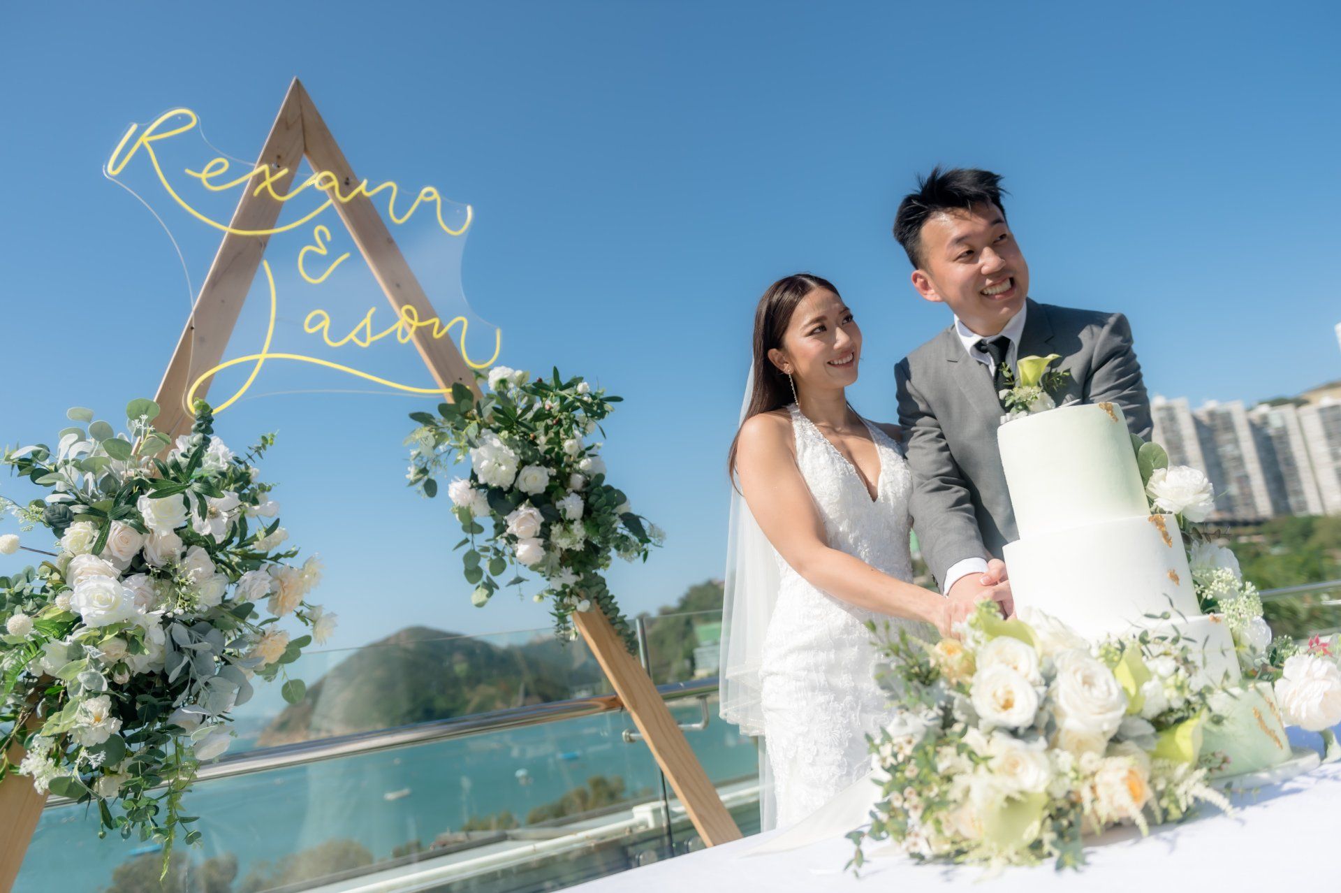 Bride and groom cutting wedding cake outdoors, sunny sky. Greenery, white cake, happy expressions.