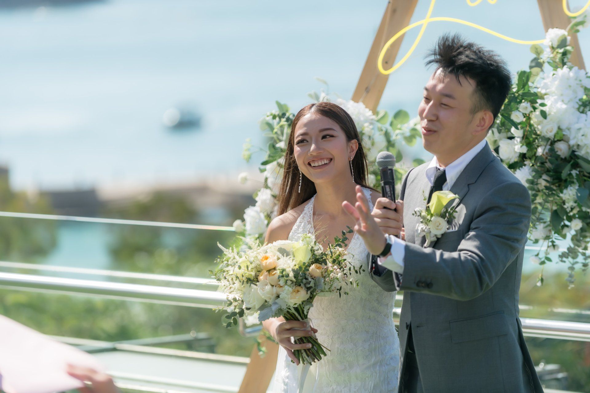 Bride and groom at outdoor wedding ceremony. Bride in white dress holds flowers, smiles. Groom speaks into microphone.