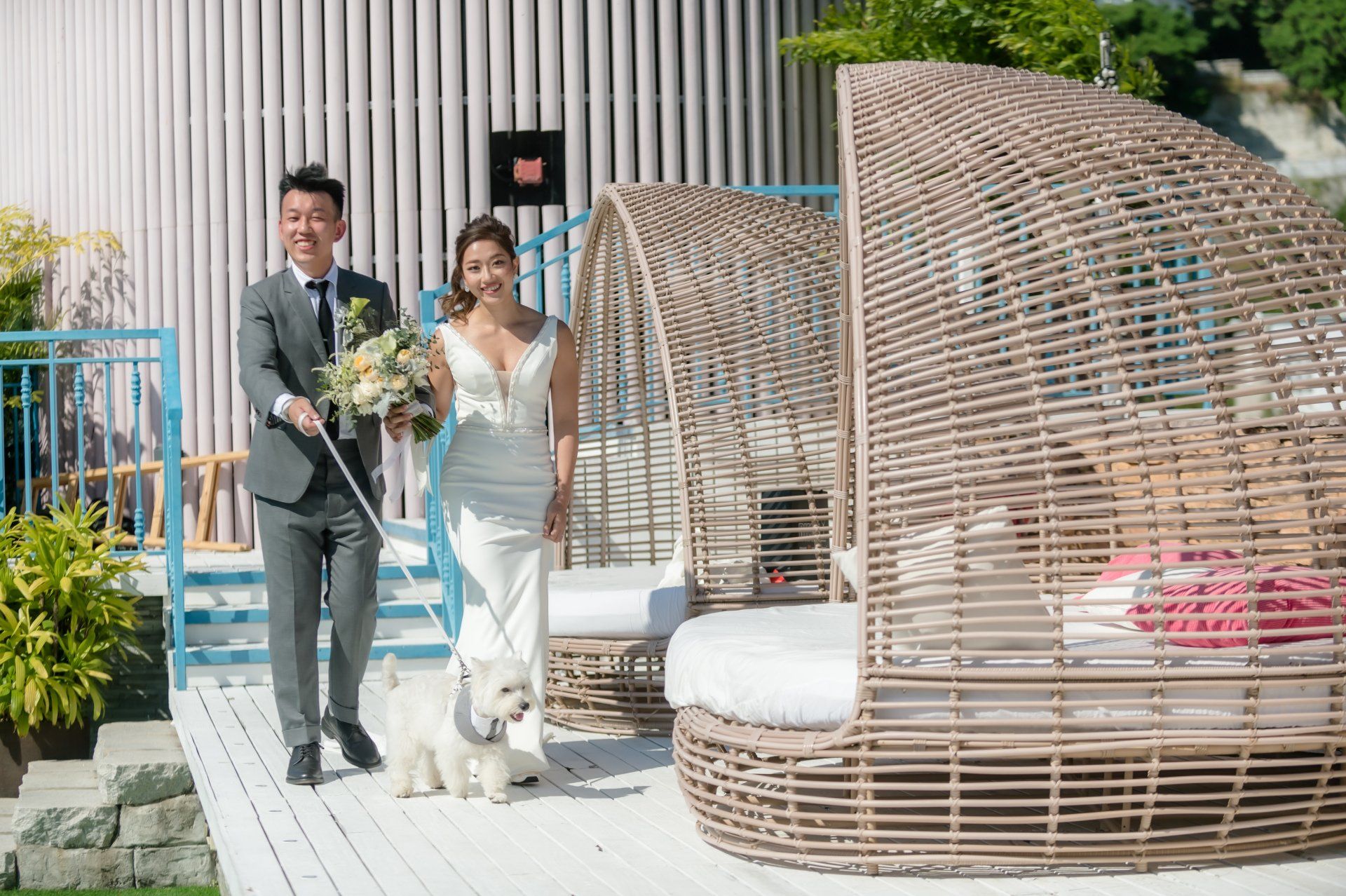 Newlyweds with a dog walking on a deck near wicker cabanas. Man holds the dog’s leash; woman carries flowers. Bright, outdoor setting.
