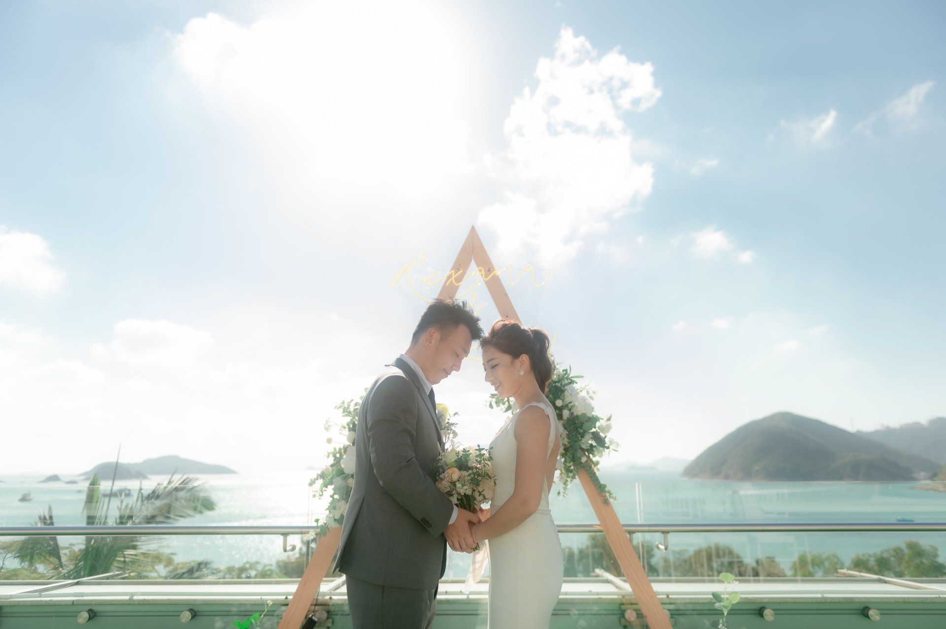Couple holding hands under a wooden arch with flowers, overlooking a sunny seascape.