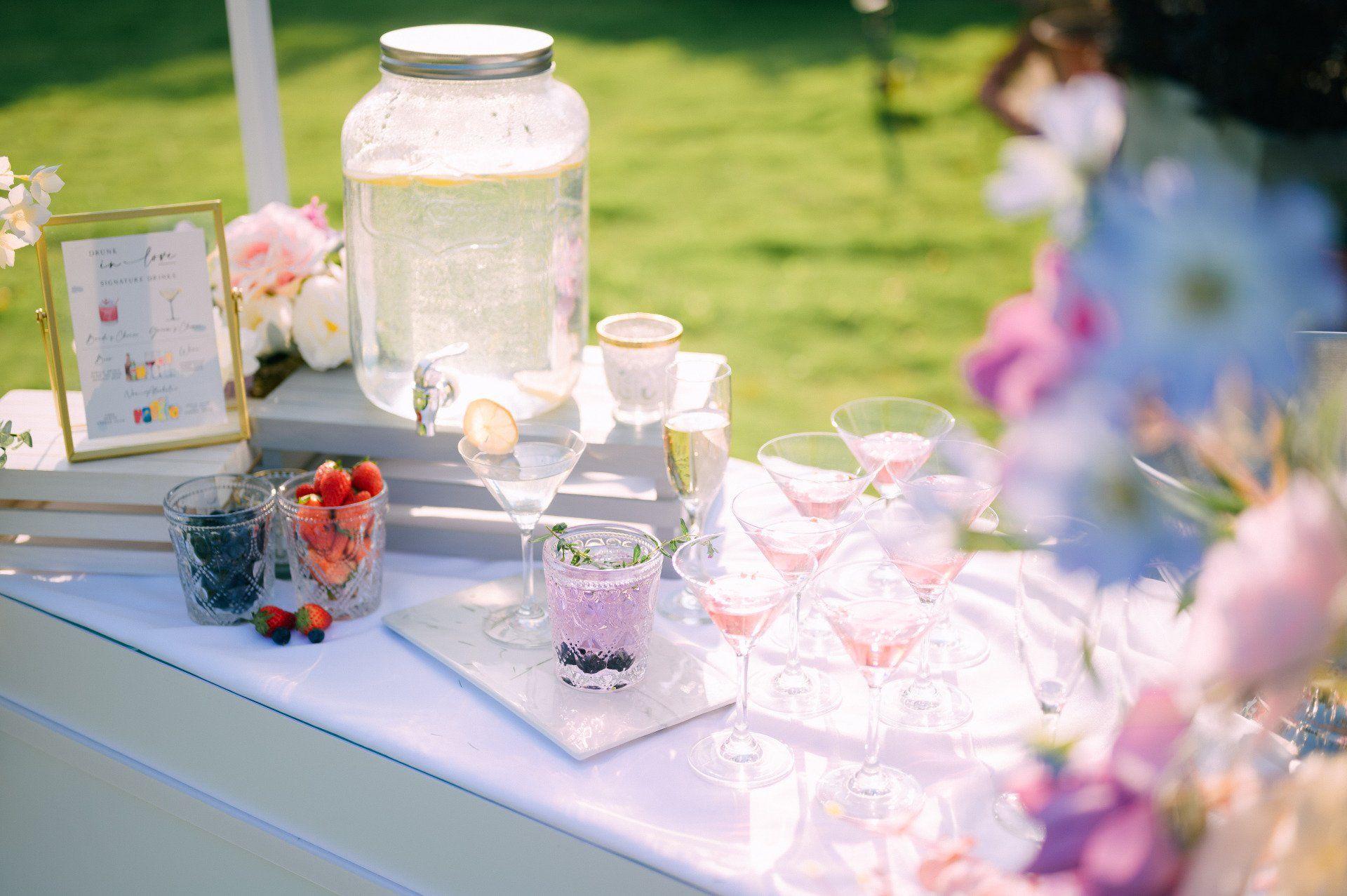 Cocktail bar set up outdoors with drinks, fruits, and flowers.