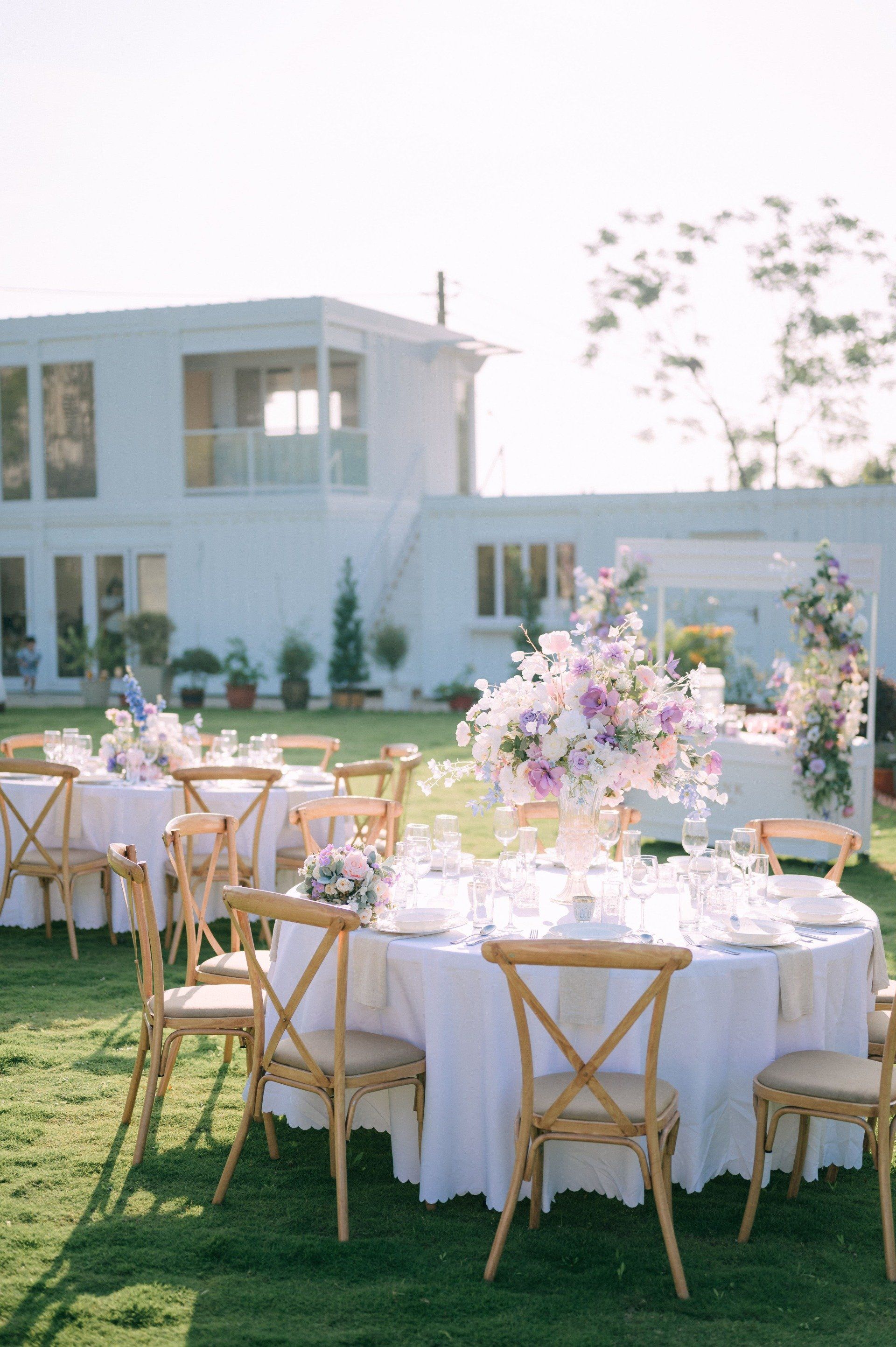 Outdoor reception tables set for dining, with floral centerpieces, on a green lawn, white building background.