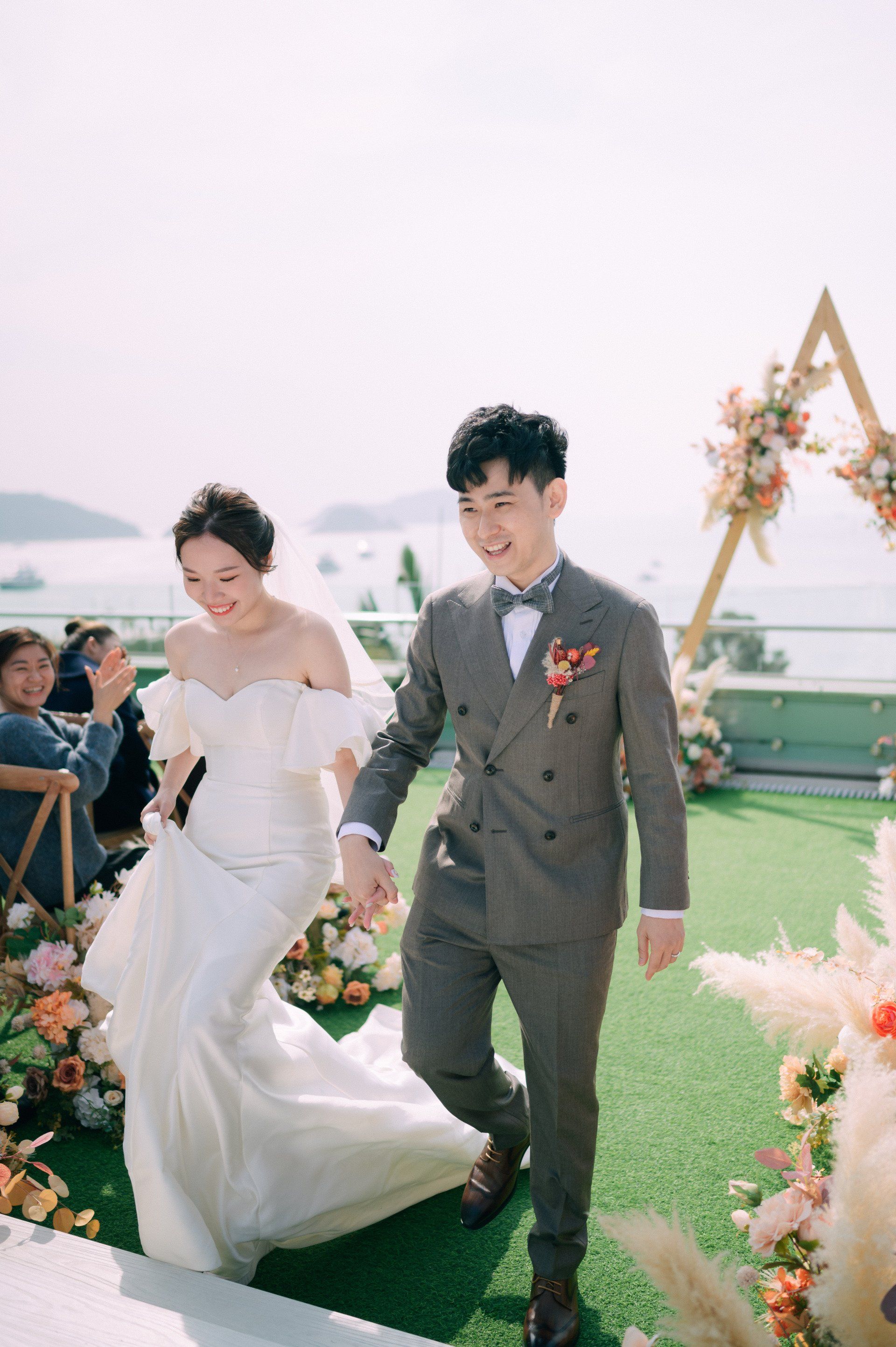 Bride and groom holding hands, exiting wedding ceremony, smiles. Outdoor rooftop with floral decor, sunny day.