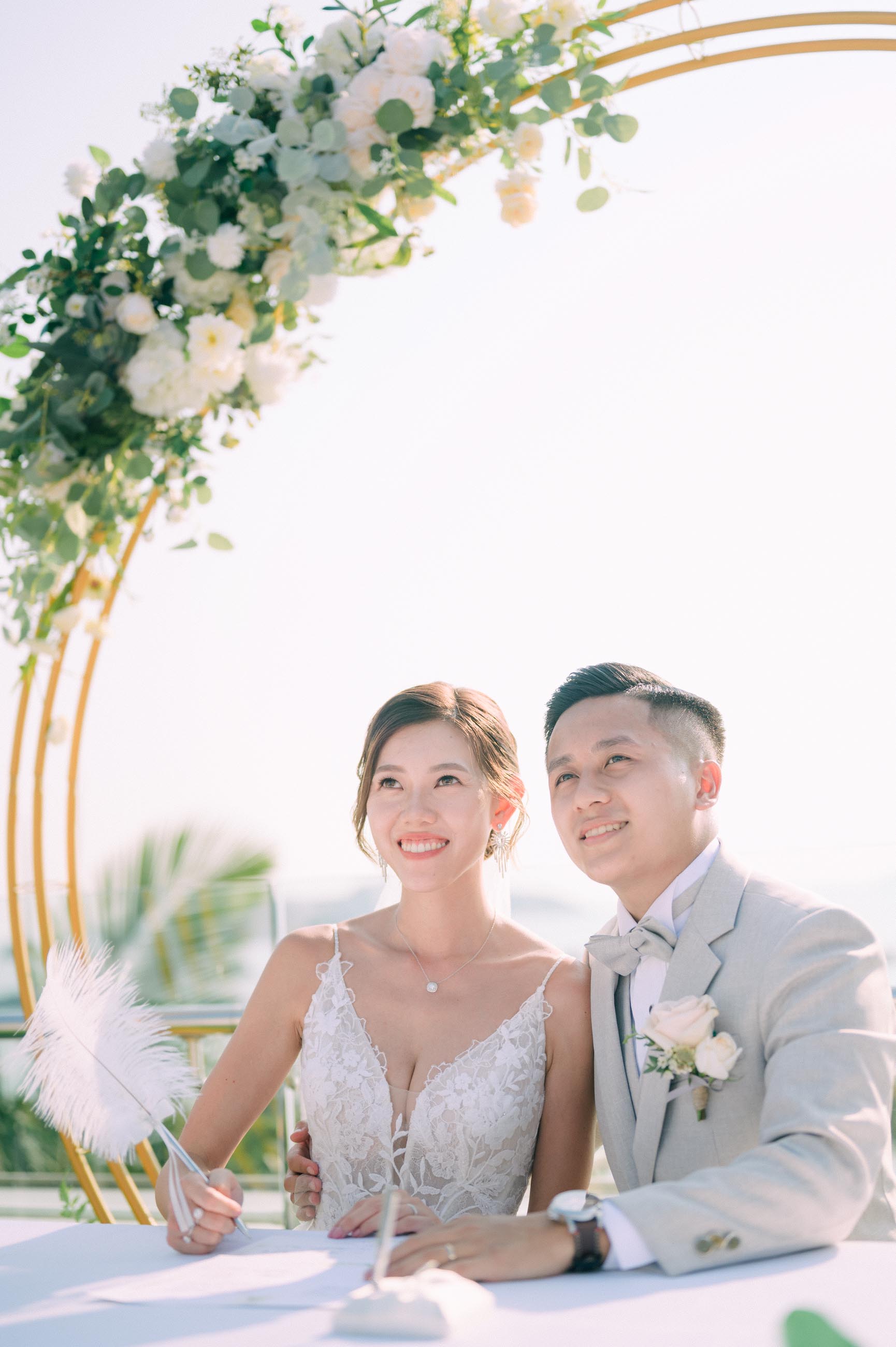 Bride and groom signing marriage documents under a floral archway.