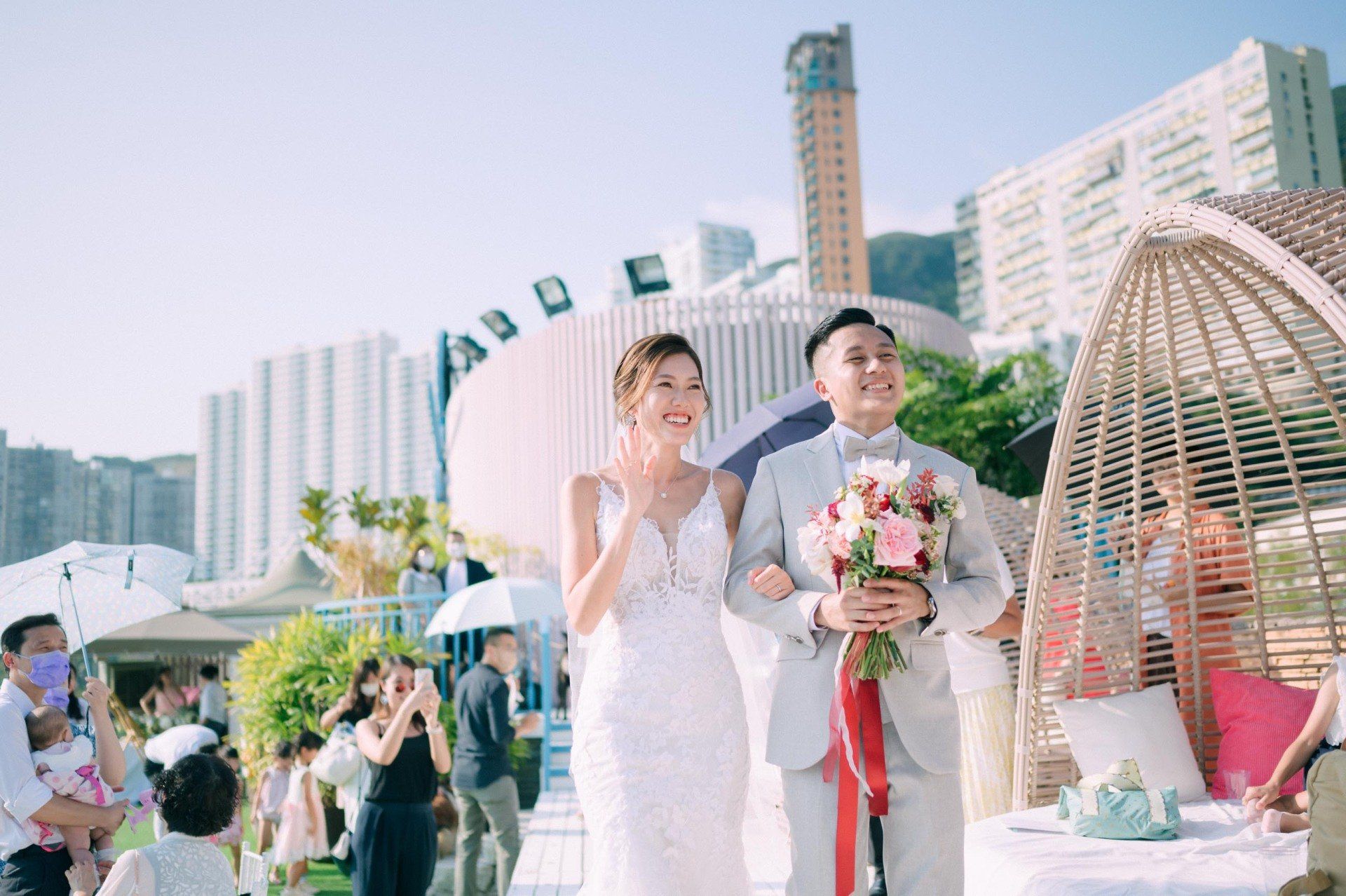 Bride and groom walk at outdoor wedding. Bride in white dress, groom in grey suit, holding flowers.