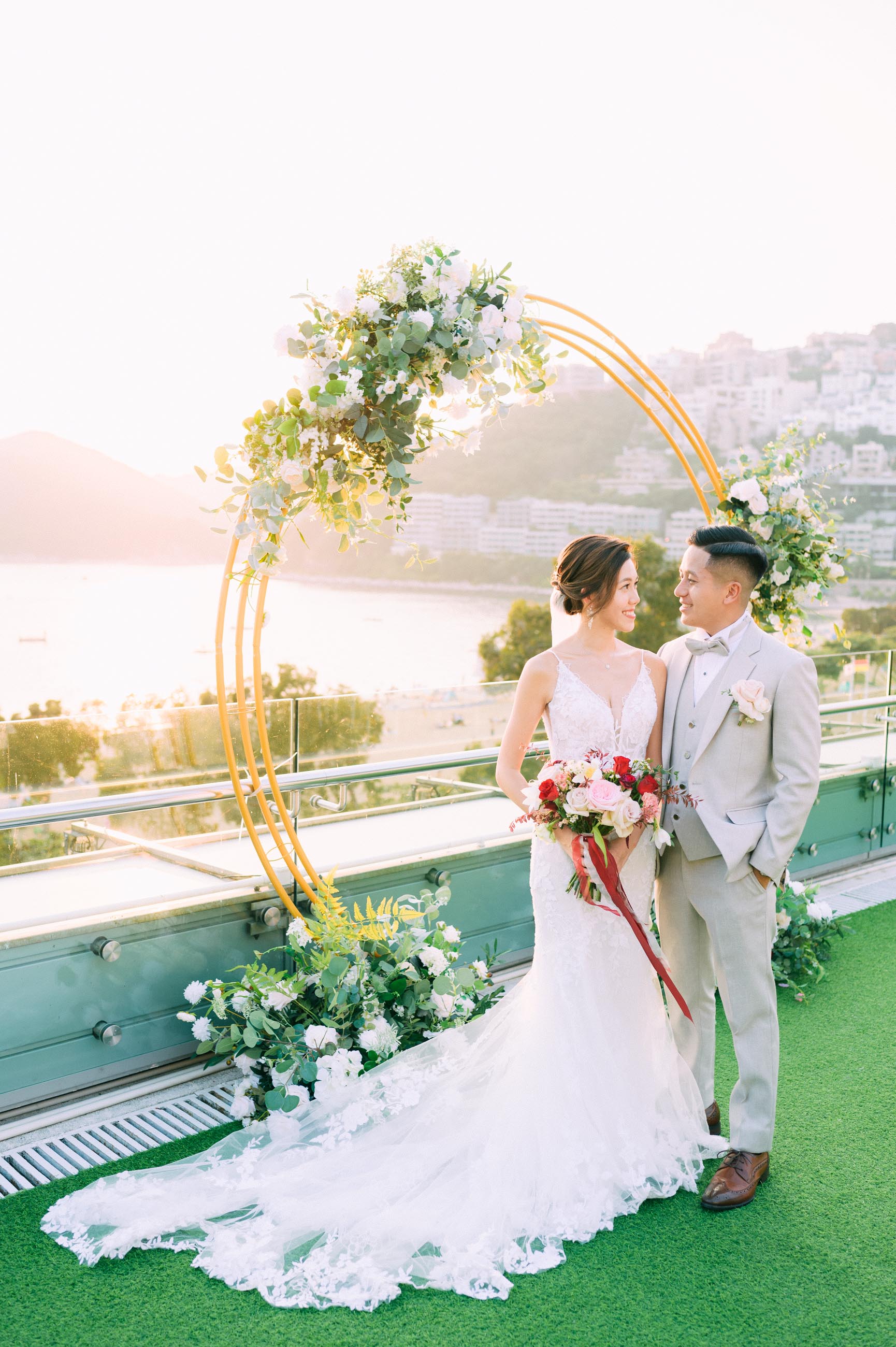 Bride and groom pose near a gold circular arch decorated with flowers, with a coastal backdrop at sunset.