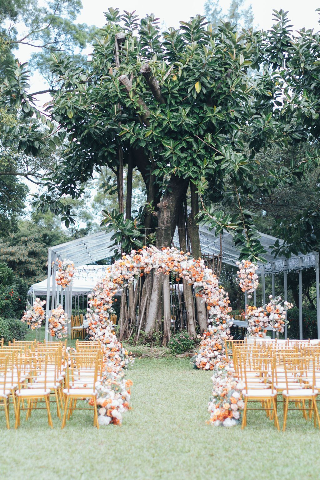 Wedding ceremony setup under a large tree with floral arch and gold chairs on the grass.