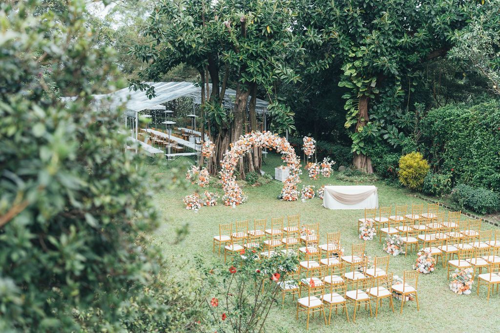 Wedding ceremony setup in a garden, rows of gold chairs facing an arch and table under a tree.