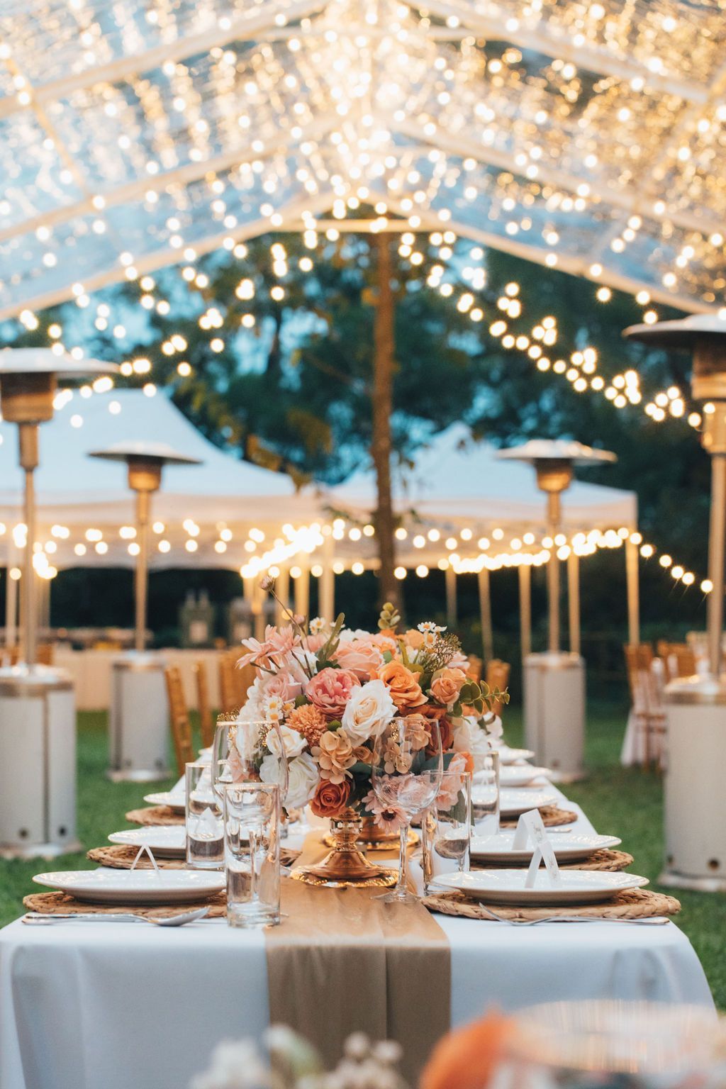 Wedding reception table with floral centerpiece under a clear tent, strung with lights, in a garden.