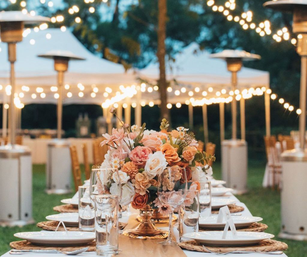 Outdoor dining table set with flowers, lit by string lights and outdoor heaters.