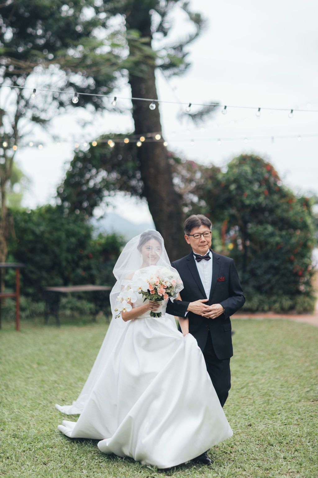 Bride walking down an outdoor aisle with her father, holding a bouquet, dressed for a wedding.