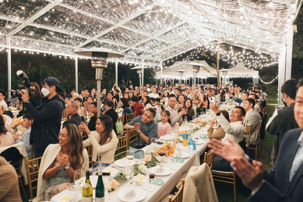 Large outdoor event under a tent, decorated with lights. People are seated at long tables, clapping and smiling.