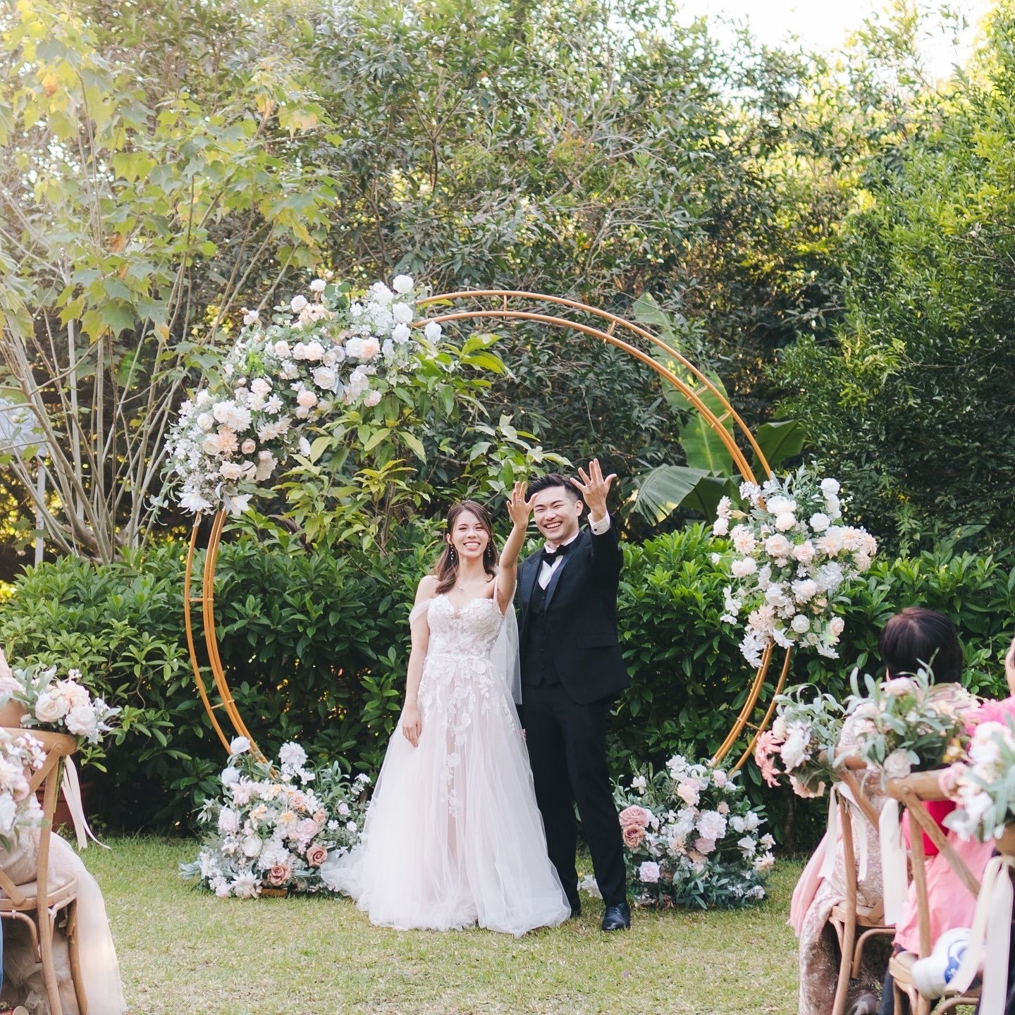 Couple at outdoor wedding ceremony, posing near golden arch decorated with white flowers.