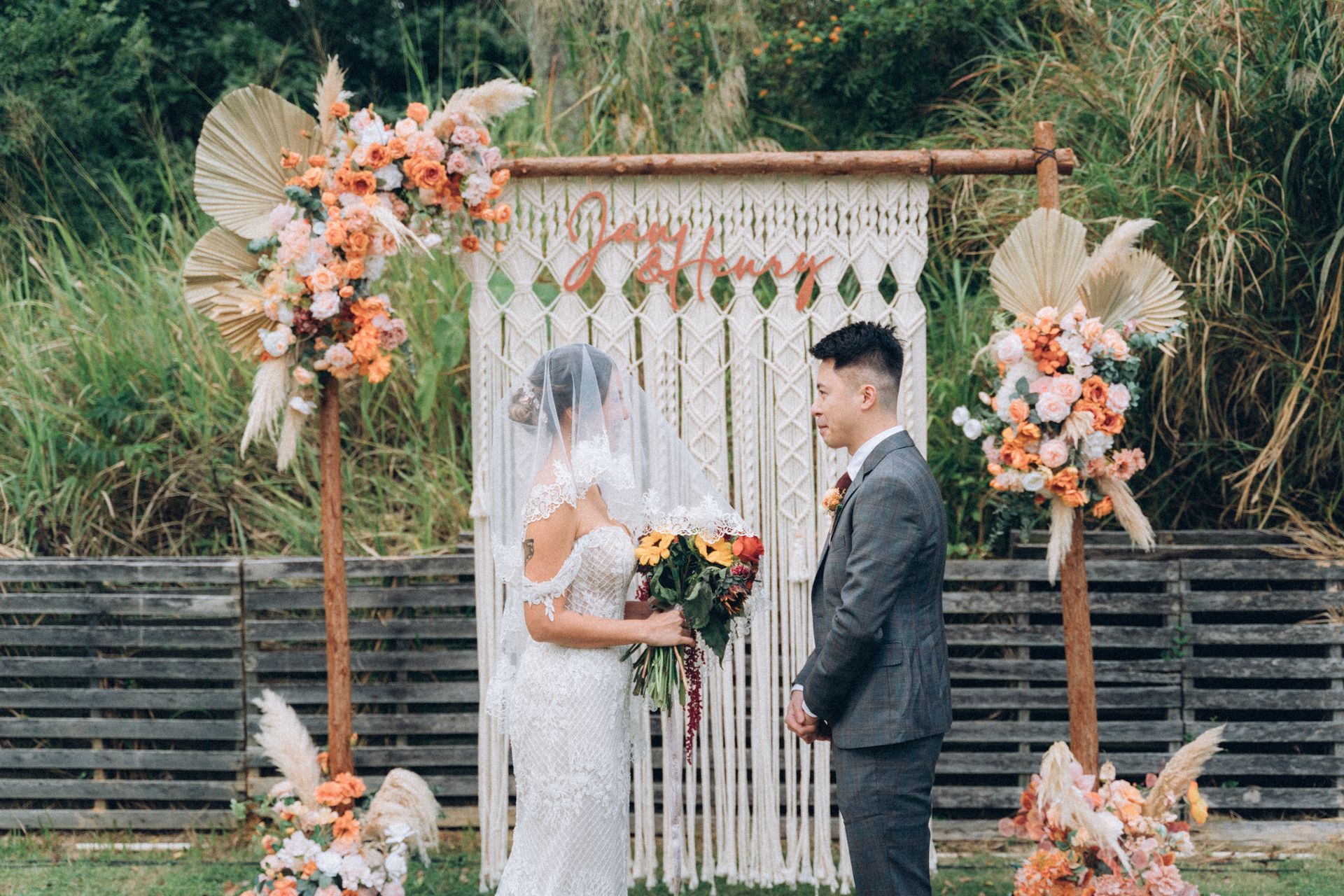 Couple at outdoor wedding ceremony; floral arch, macrame backdrop, neutral colors.