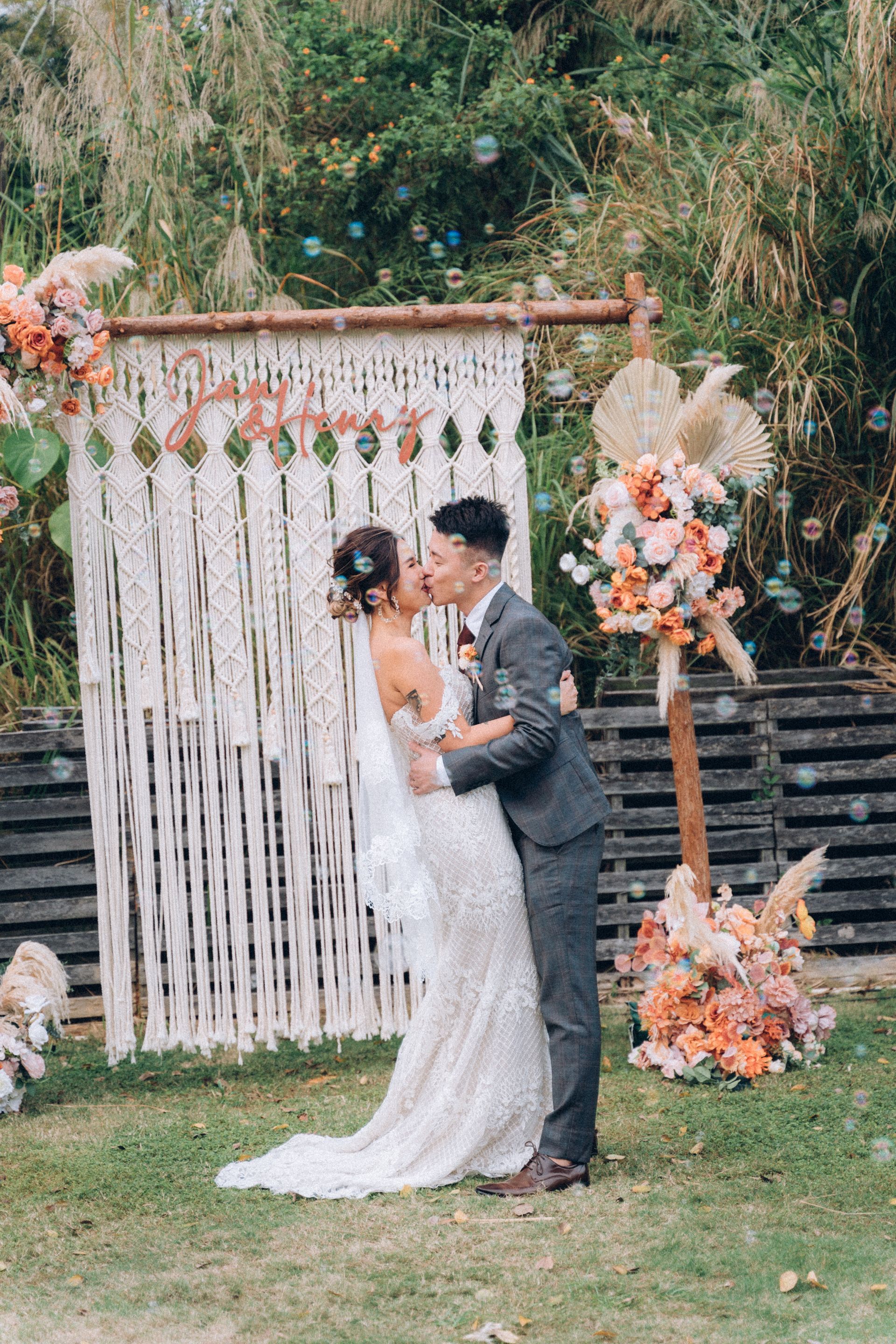 Couple kissing at outdoor wedding ceremony; floral arch, macrame backdrop, and bubbles.