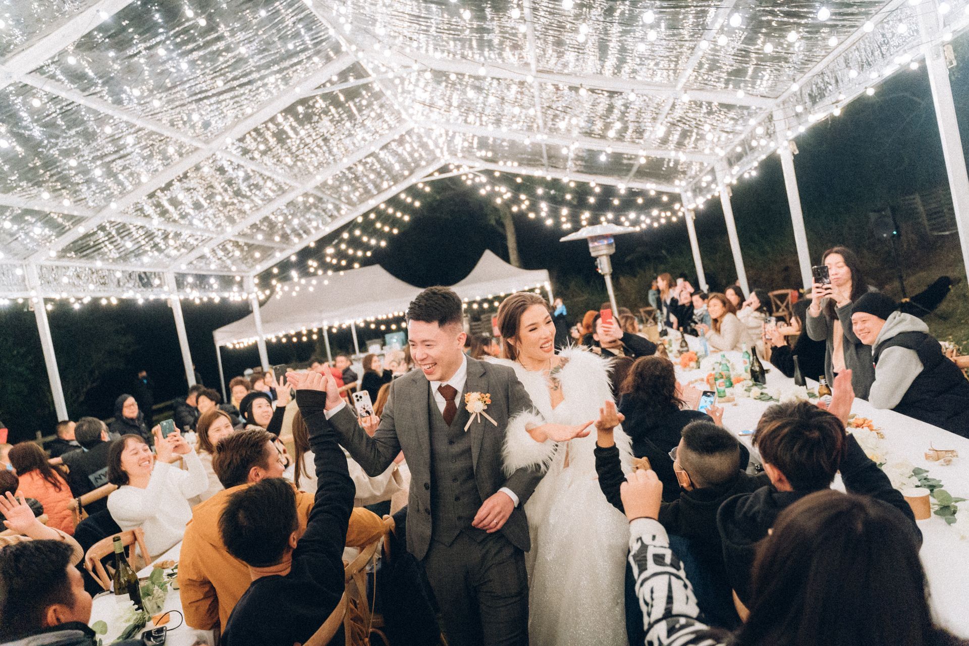 Newly married couple celebrates under a canopy of lights with cheering guests.