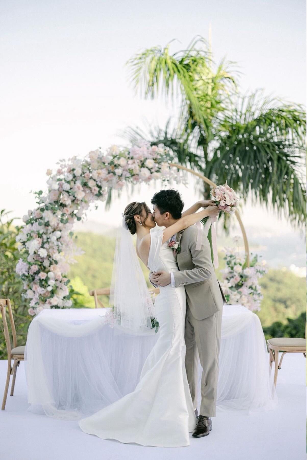 Bride and groom kissing under a floral arch at an outdoor wedding ceremony.