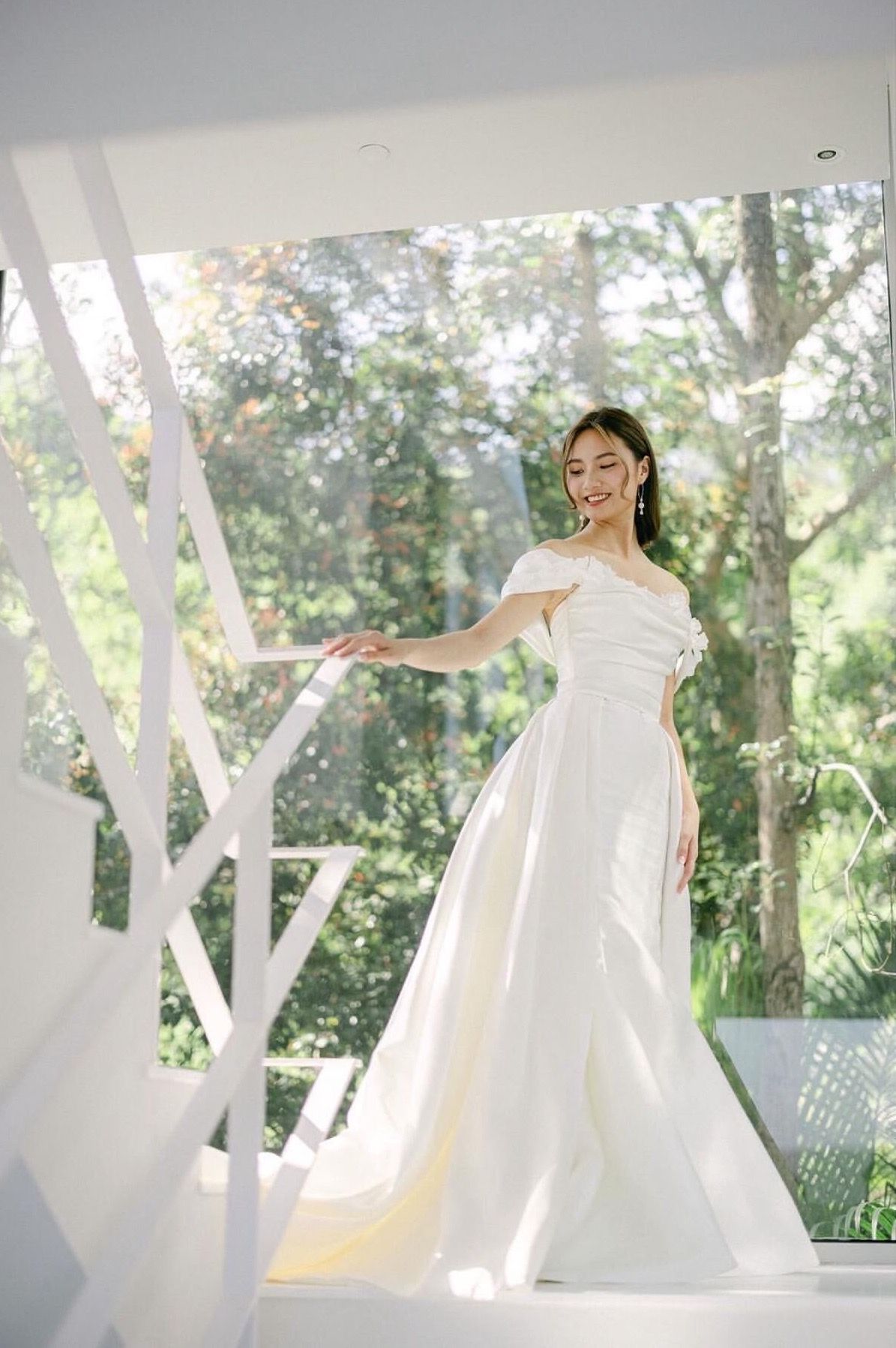 Woman in a white off-the-shoulder wedding dress stands by a modern white staircase, with a sunlit background of trees.