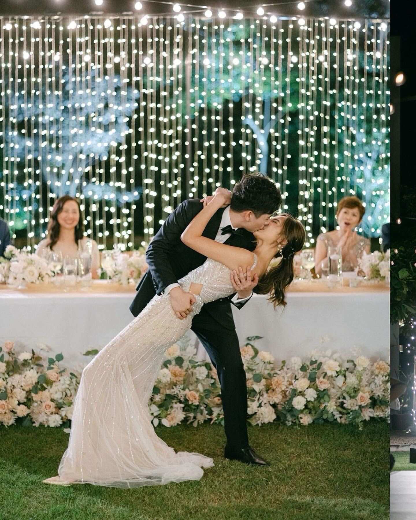 Couple kisses during a wedding reception, bending backward. String lights, floral decorations, and guests in background.