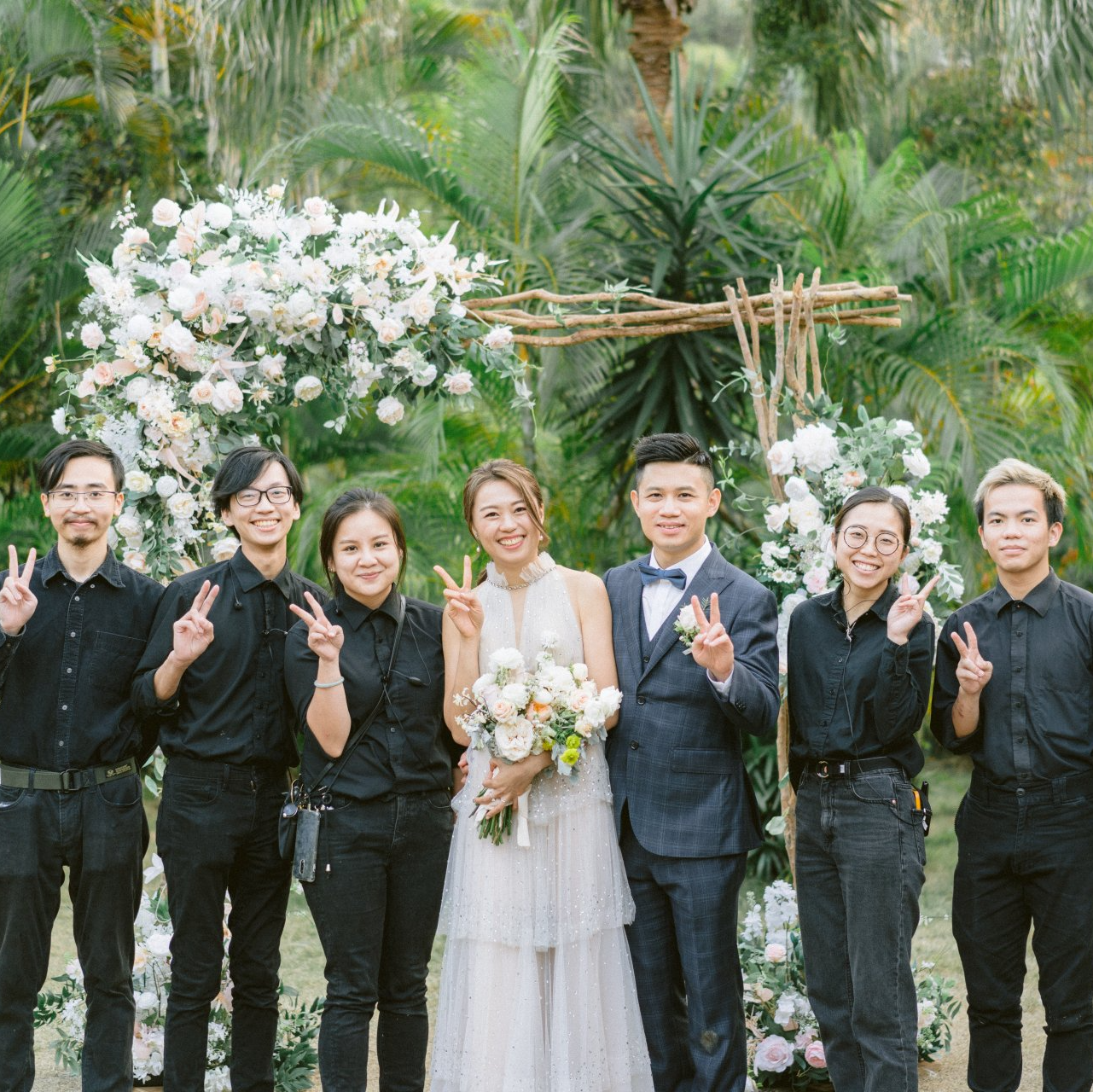 Bride and groom with wedding crew, posed in front of floral arch. Everyone is giving peace signs.