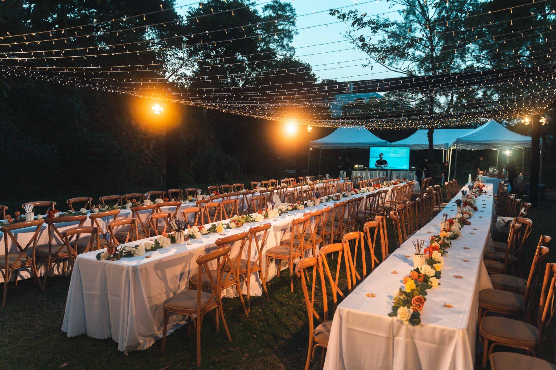 Outdoor reception with long tables, white tablecloths, floral centerpieces, string lights.